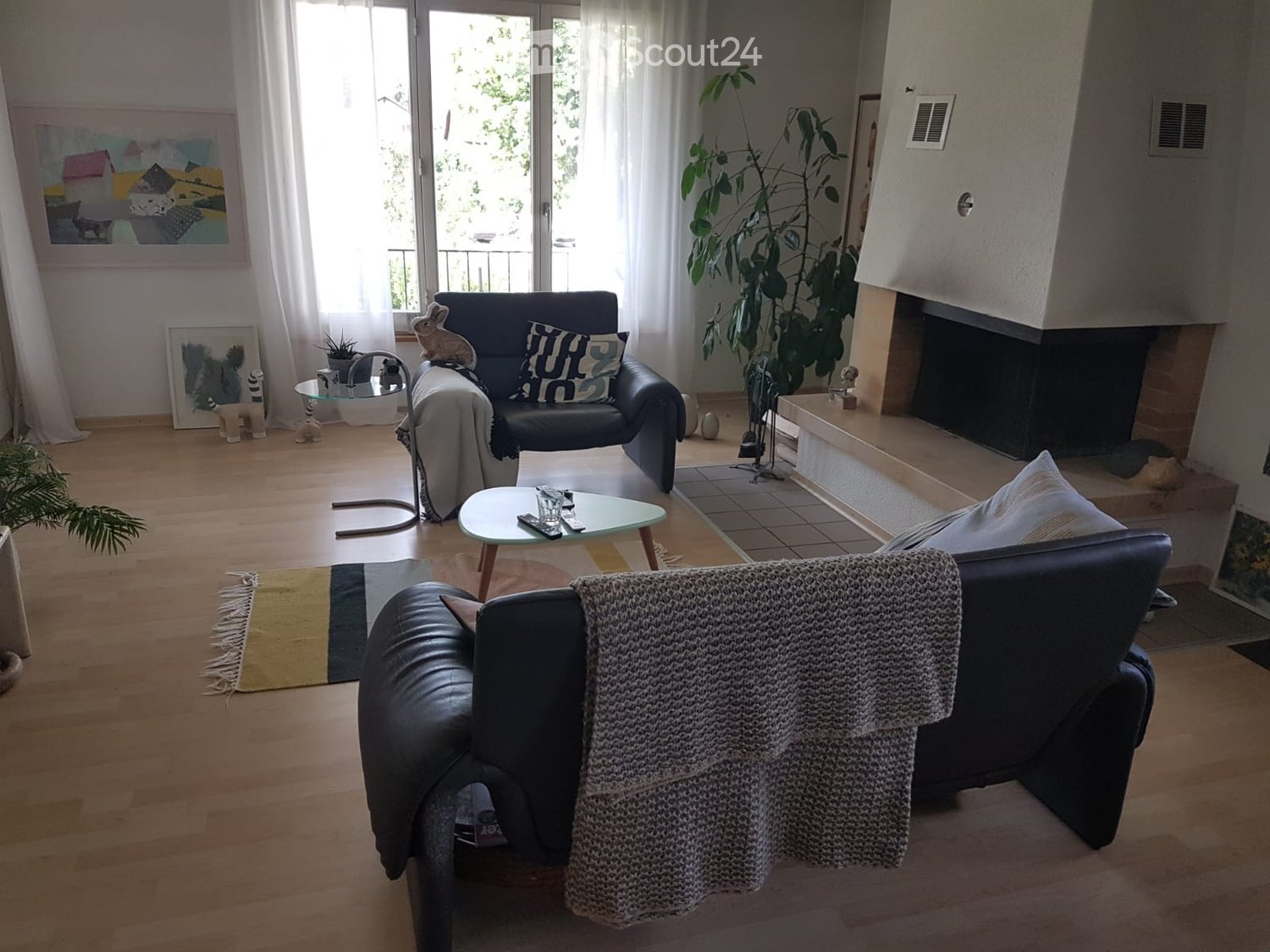Modern living room with hardwood floors, fireplace, large windows, balcony access, sofa, coffee table, armchairs, ottoman, floor lamp, and decorative items.