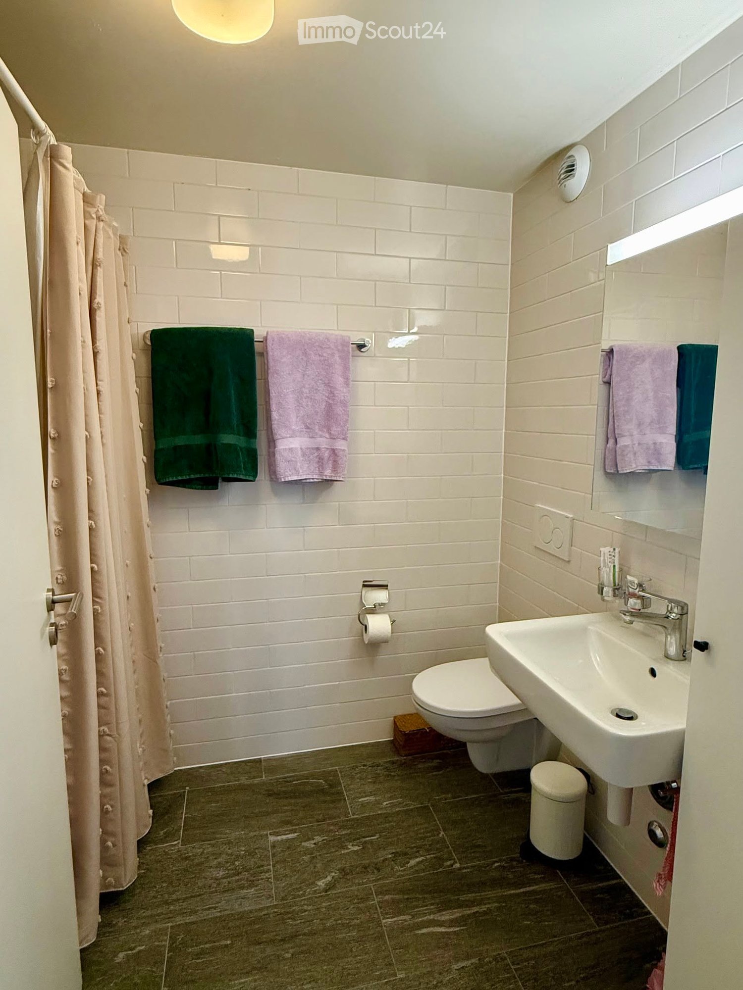 Modern bathroom with white tiled walls, dark grey tiled floor, toilet, sink, mirror, and towels.