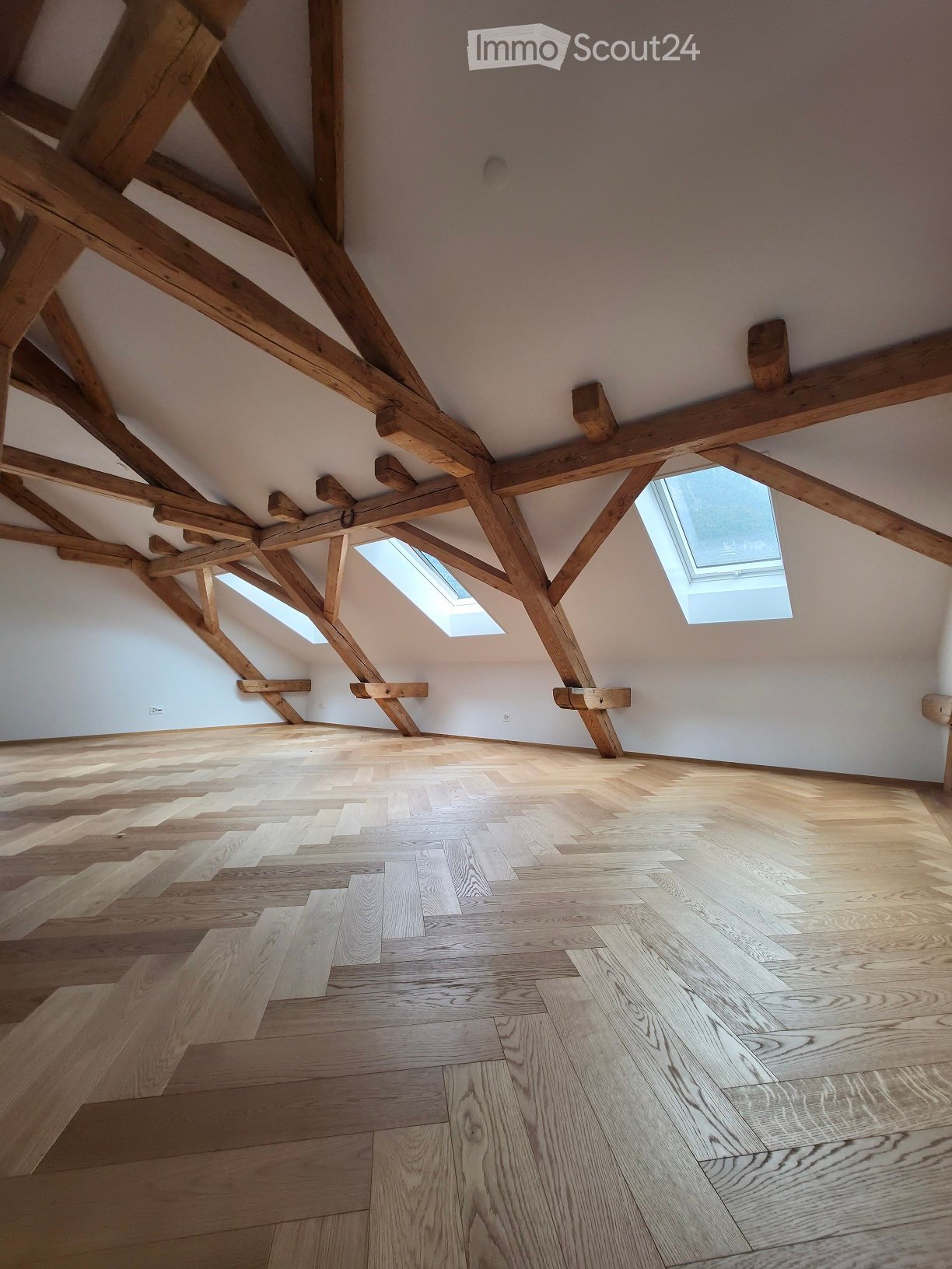 Empty attic space with exposed wooden beams, light-colored wooden flooring, and skylight windows allowing natural light.