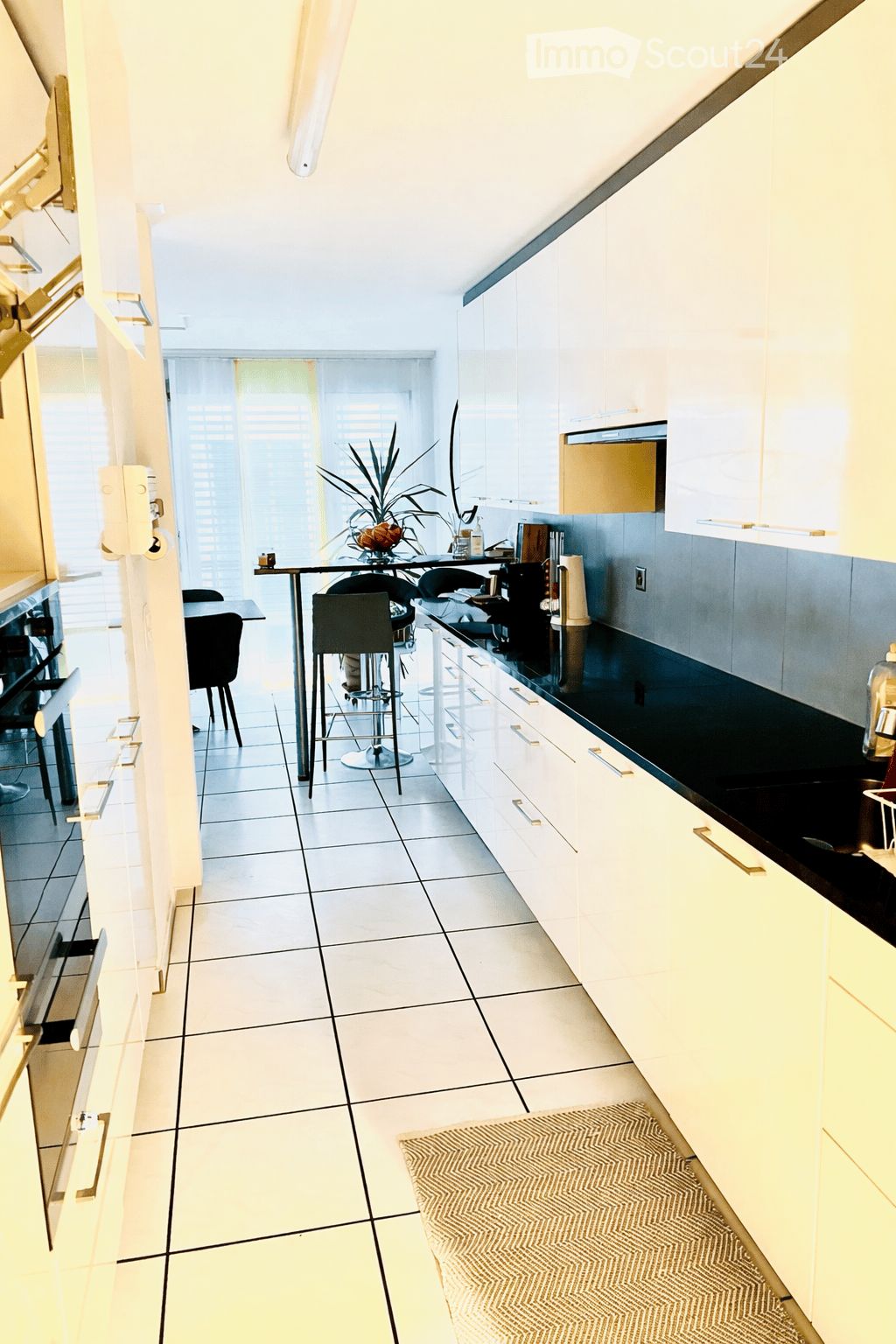 Modern kitchen with white cabinets, black countertops, and a dining area with a table and stools. Features include a built-in oven, dishwasher, and a small plant on the counter.