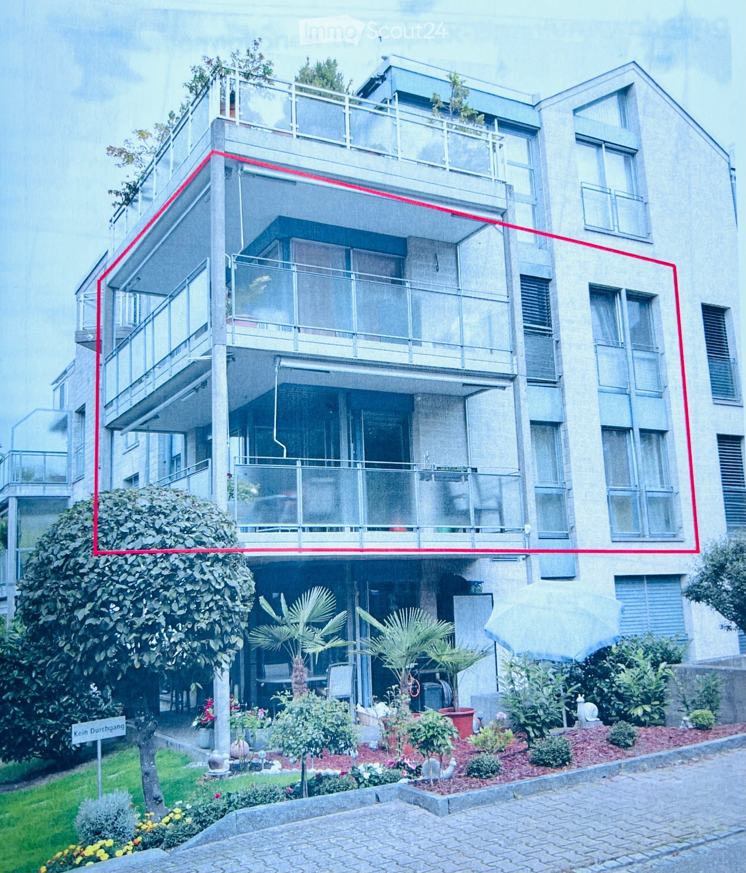 Modern multi-story residential building with balconies, glass railings, and a landscaped garden area with palm trees and shrubs.