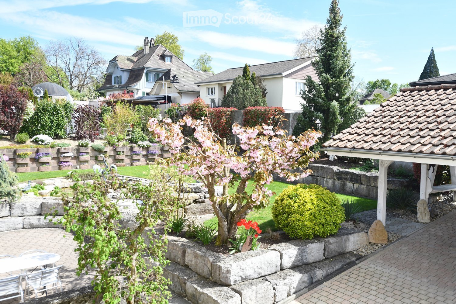 Well-maintained garden with a stone wall, flowering trees, shrubs, and a patio area with outdoor furniture.