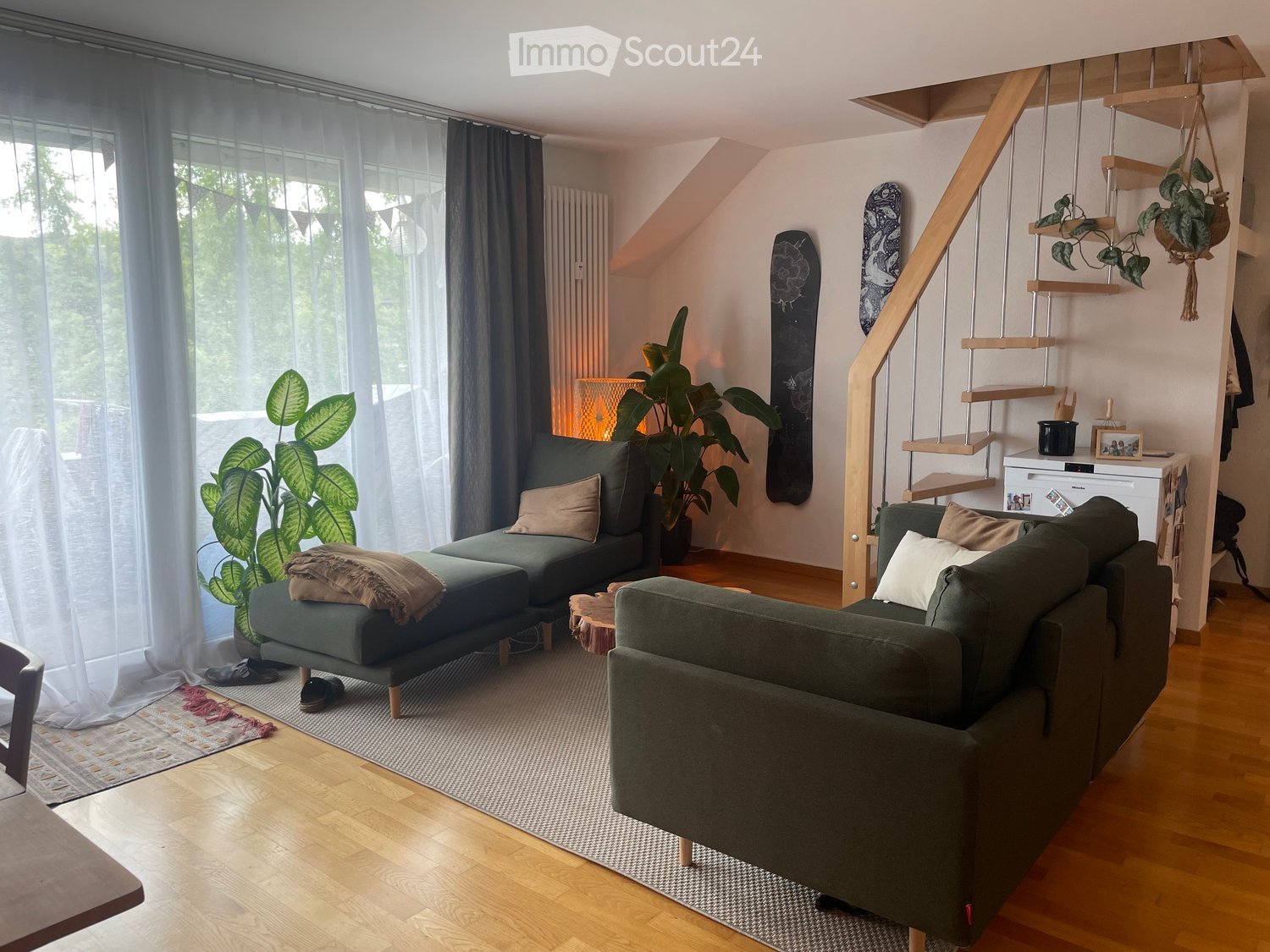 Modern living room with wooden flooring, large windows, green sofa, ottoman, small table, hanging plants, and skateboards on the wall.