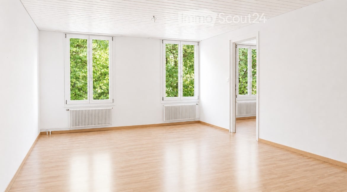 Empty room with white walls, wooden floor, large windows with green views, radiators, and a doorway leading to another space.