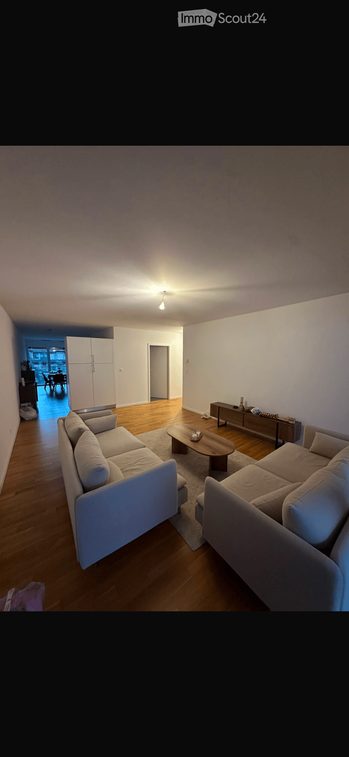 Modern living room with light-colored wooden flooring, a large sectional sofa, a coffee table, and a side table with decorative items. The room features white walls and a ceiling light fixture.