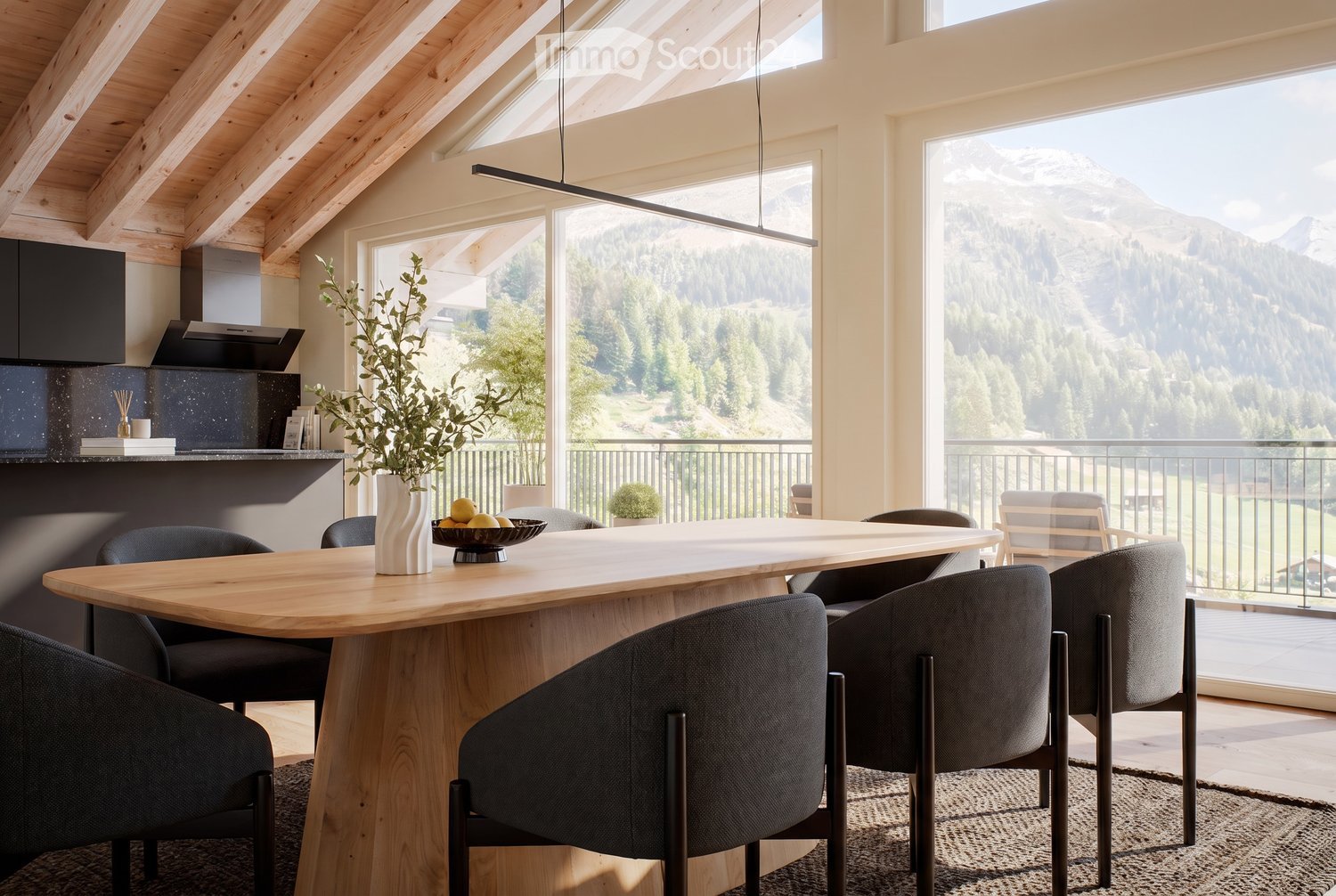 Modern dining area with a large wooden table, grey upholstered chairs, a vase with greenery, and a bowl of fruit. The space features exposed wooden beams, large windows with a scenic mountain view, and contemporary kitchen elements.