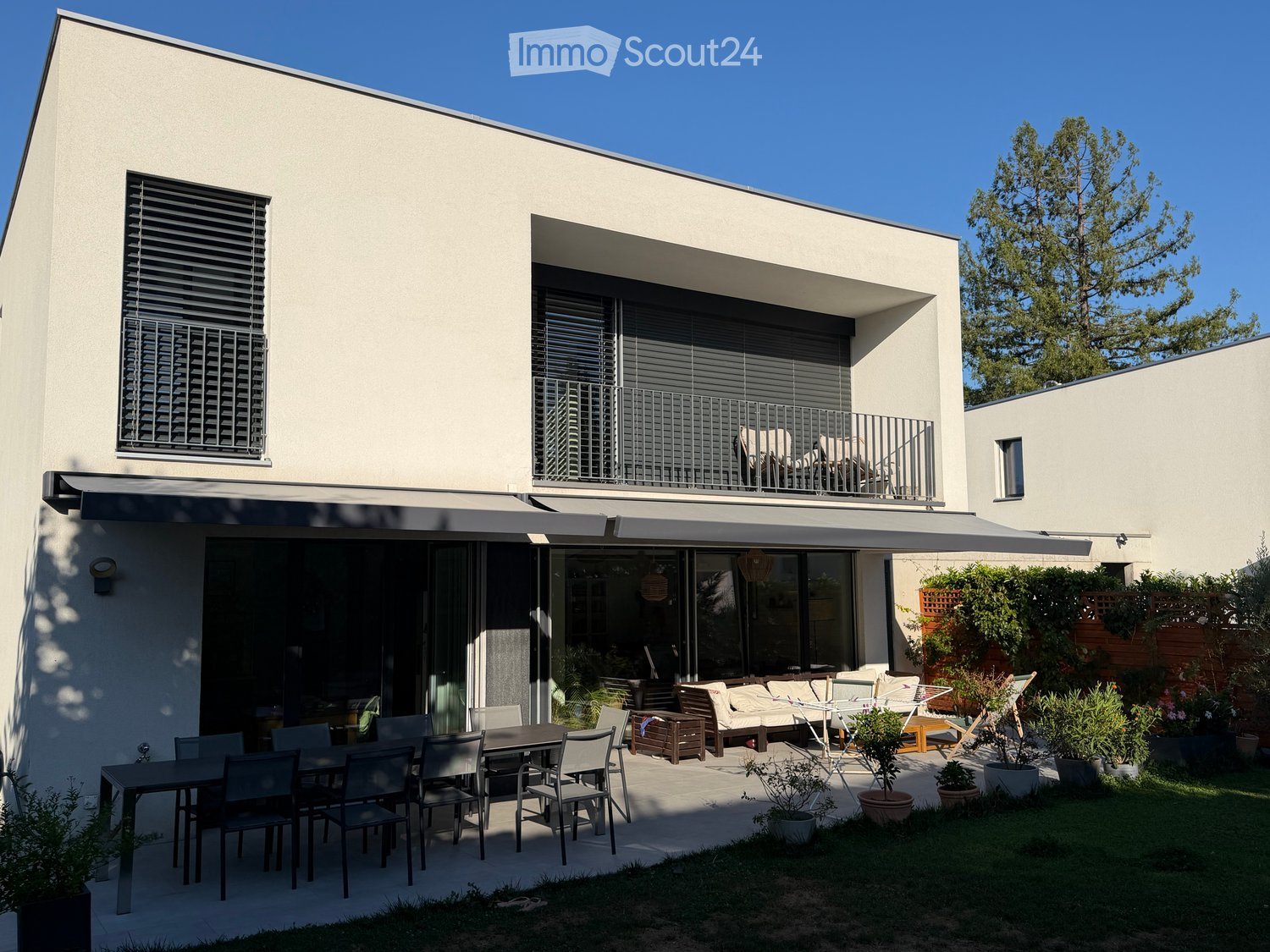 Modern house exterior with a spacious patio area featuring a dining table, chairs, a sofa, and potted plants. The house has large windows and a balcony with additional seating.