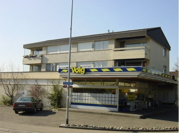 Multi-story building with balconies, storefront signage reading "Volg", and a parked car in front.