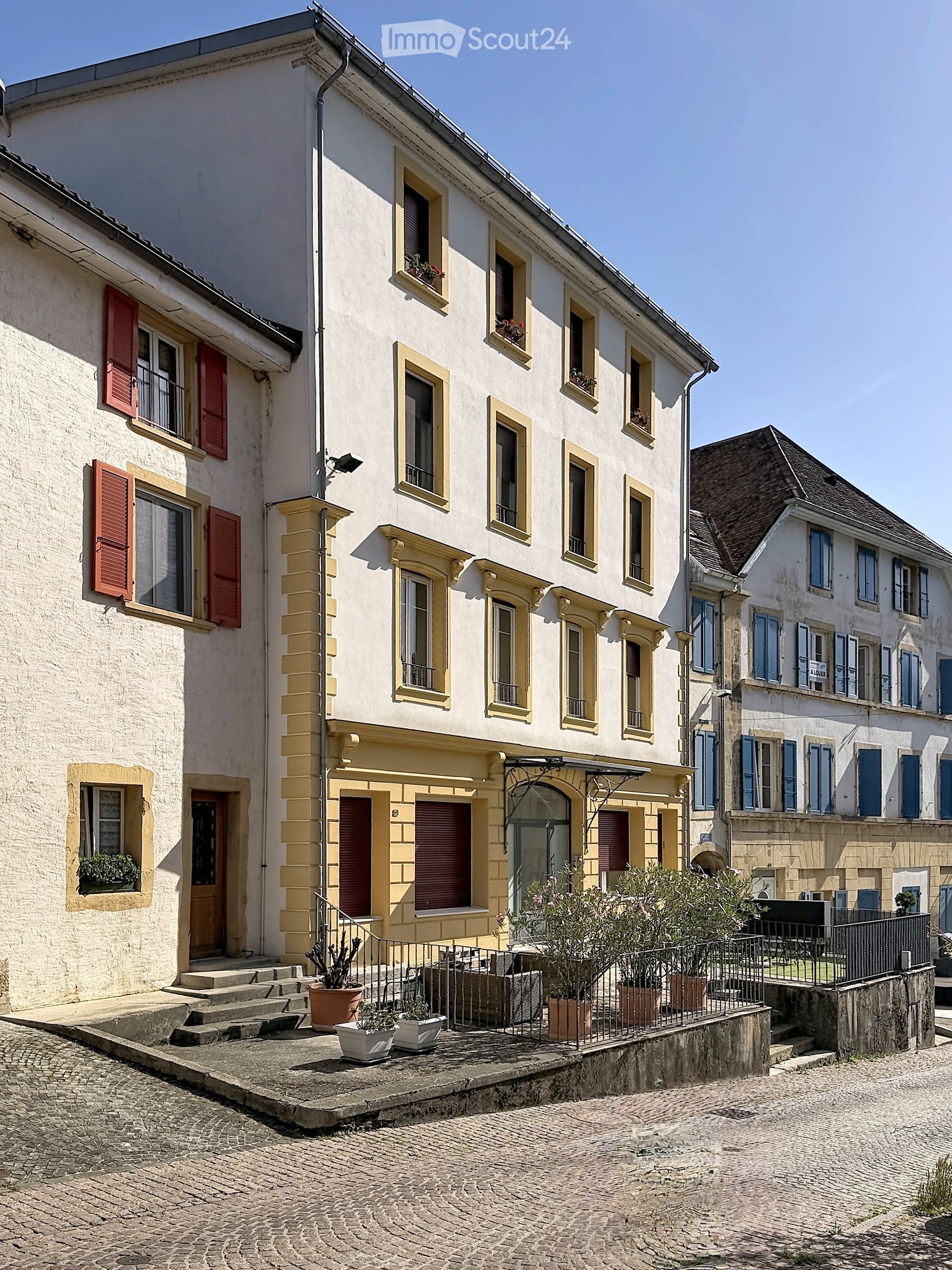 A multi-story building with white walls, red shutters, and multiple windows, featuring potted plants on the raised ground floor.