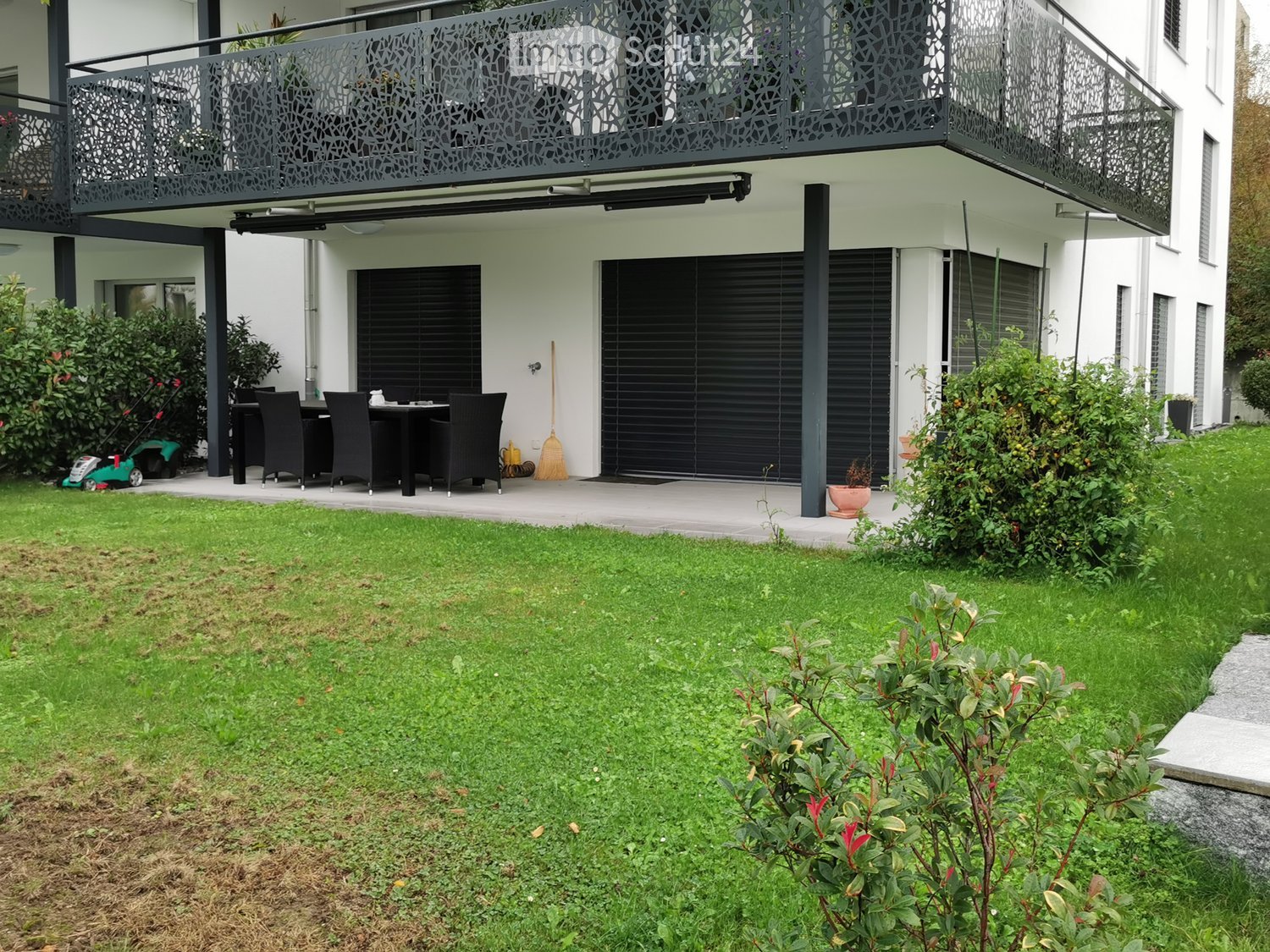 Well-maintained garden with green lawn, small patio area with a table and chairs, and a modern building with a balcony in the background.