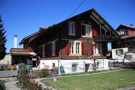 Traditional Swiss house with wooden facade, red shutters, and a small balcony. The house has multiple windows and a stone base, surrounded by a well-maintained garden with a fence and a few plants.