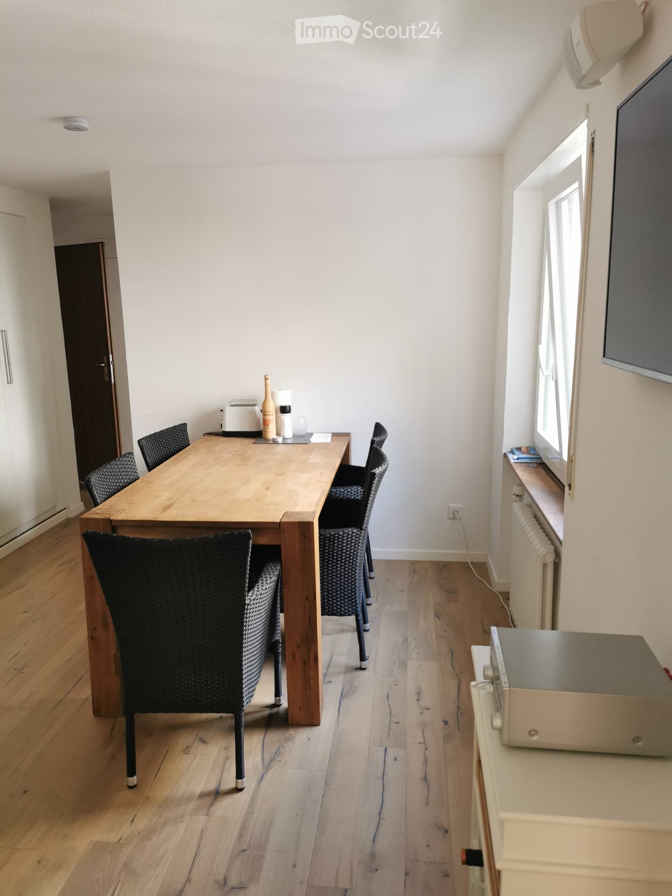A modern dining room with a wooden table, four black chairs, a small oven, and a screen mounted on the wall. The room features light wooden flooring and white walls.