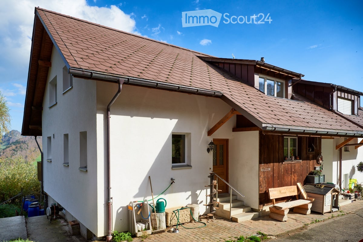 Two-story house with white walls, brown roof, wooden door, and small porch area with bench and tools.