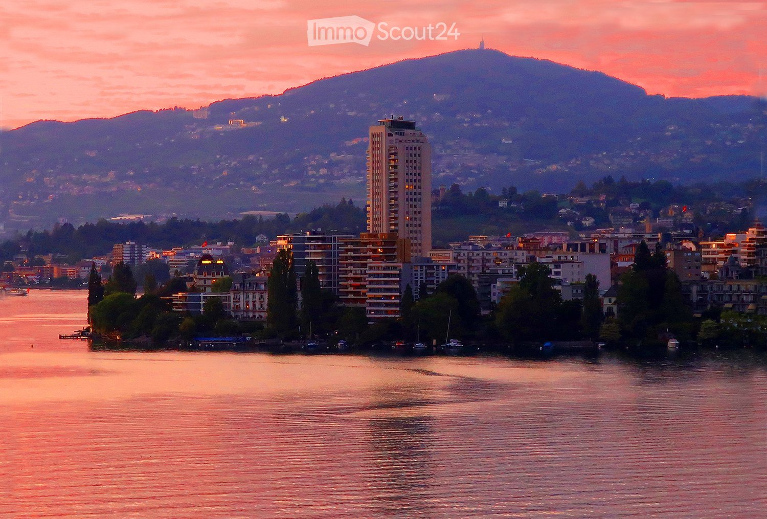 A scenic view of a cityscape at sunset, featuring a body of water in the foreground, with buildings and trees lining the shore, and mountains in the background.