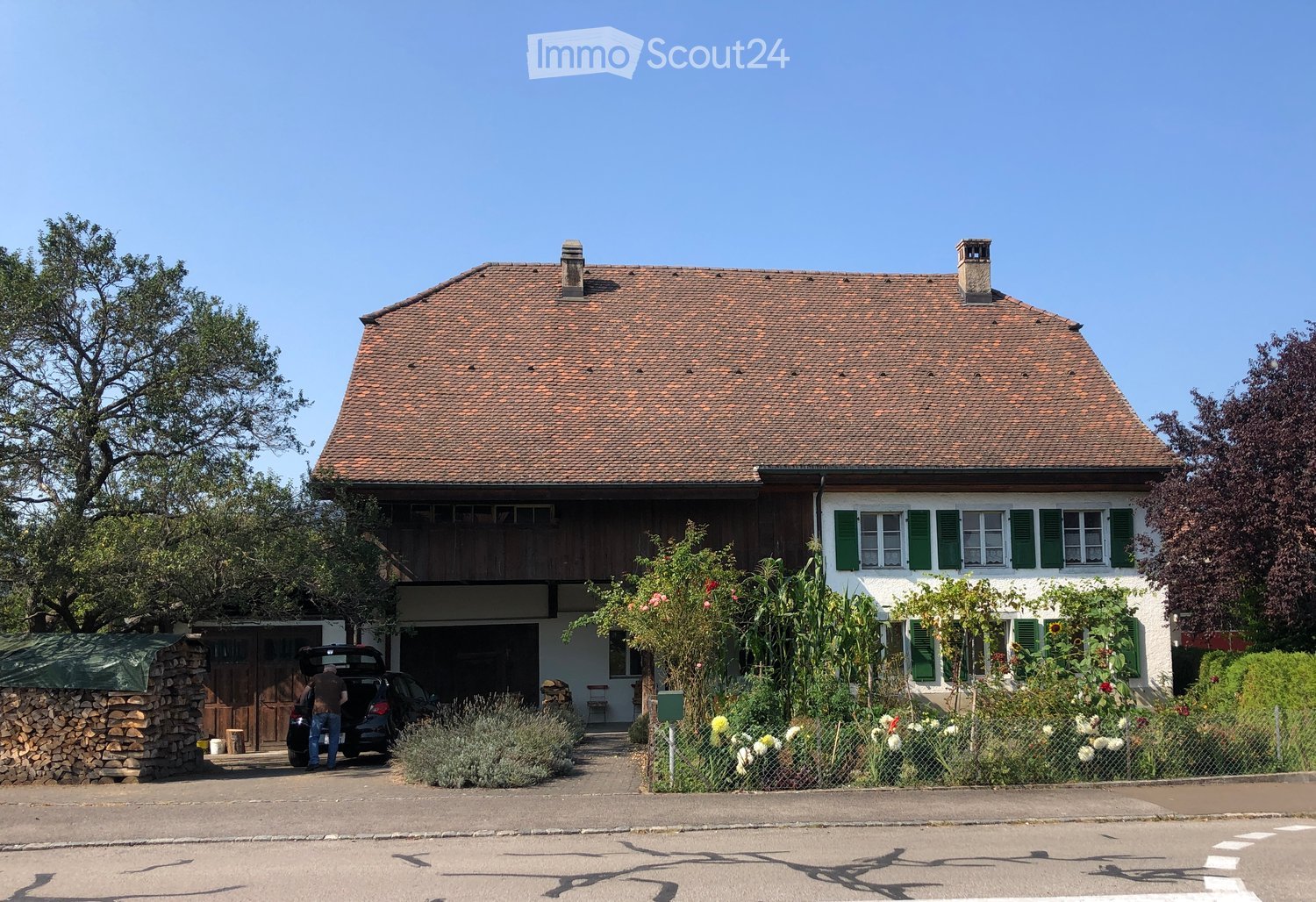 Two-story house with a red-tiled roof, white walls, green shutters, and a wooden balcony. Features a garage, garden with flowers, and a stone woodpile.
