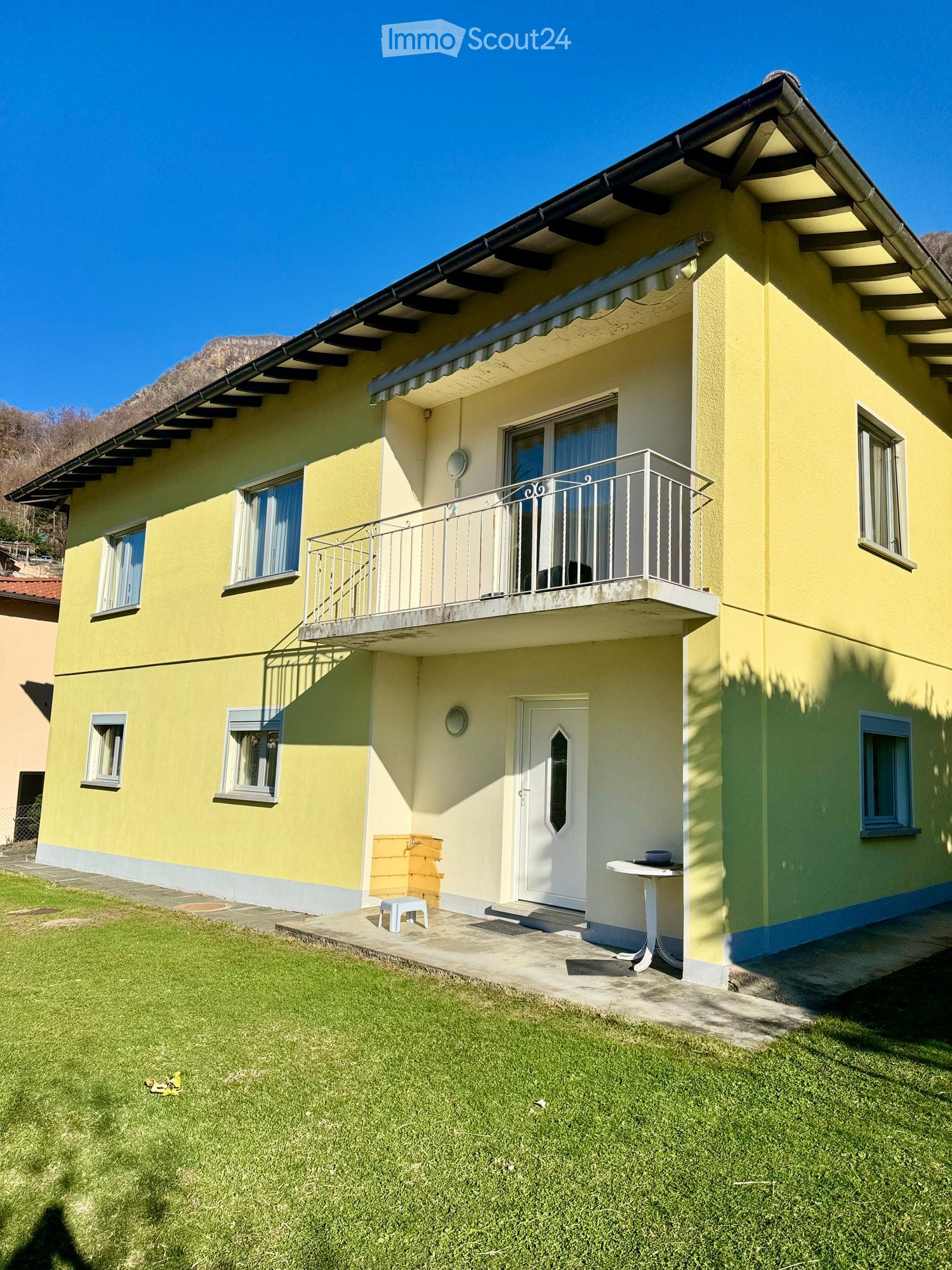Two-story yellow house with white trim, multiple windows, a balcony on the upper floor, and a small patio area with a table and chairs.