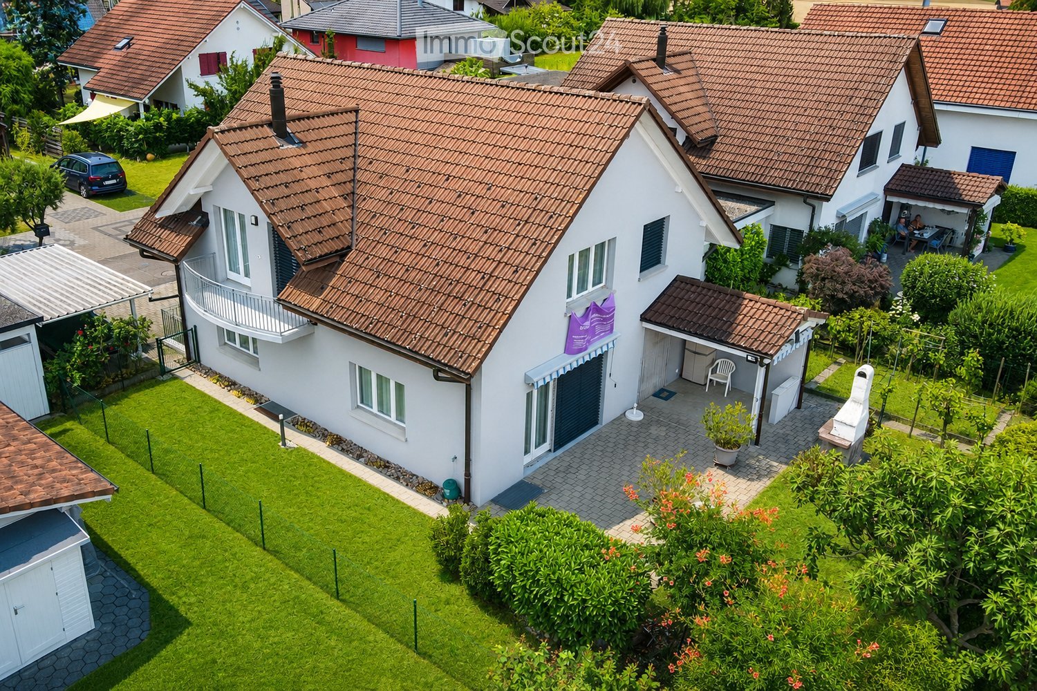 A two-story house with a red-tiled roof, white exterior, and a well-maintained garden with a barbecue and seating area.