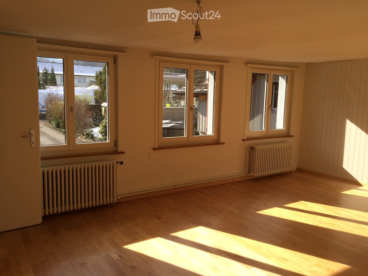 Empty room with wooden flooring, large windows, radiators, and natural light streaming in.