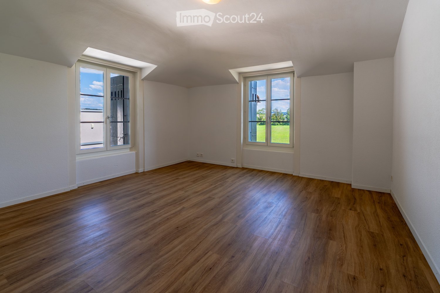 Empty room with wooden flooring, white walls, and large windows offering a view of the outdoors.