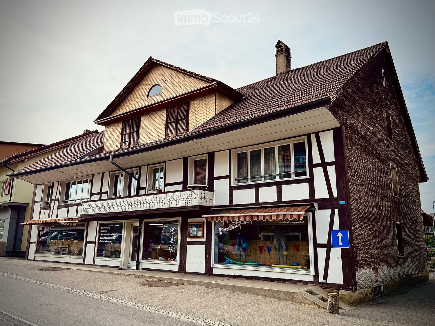 Two-story building with a tiled roof, multiple windows, and a storefront with awnings and signage. The ground floor has glass windows displaying various items, and the upper floor features balconies and decorative railings.