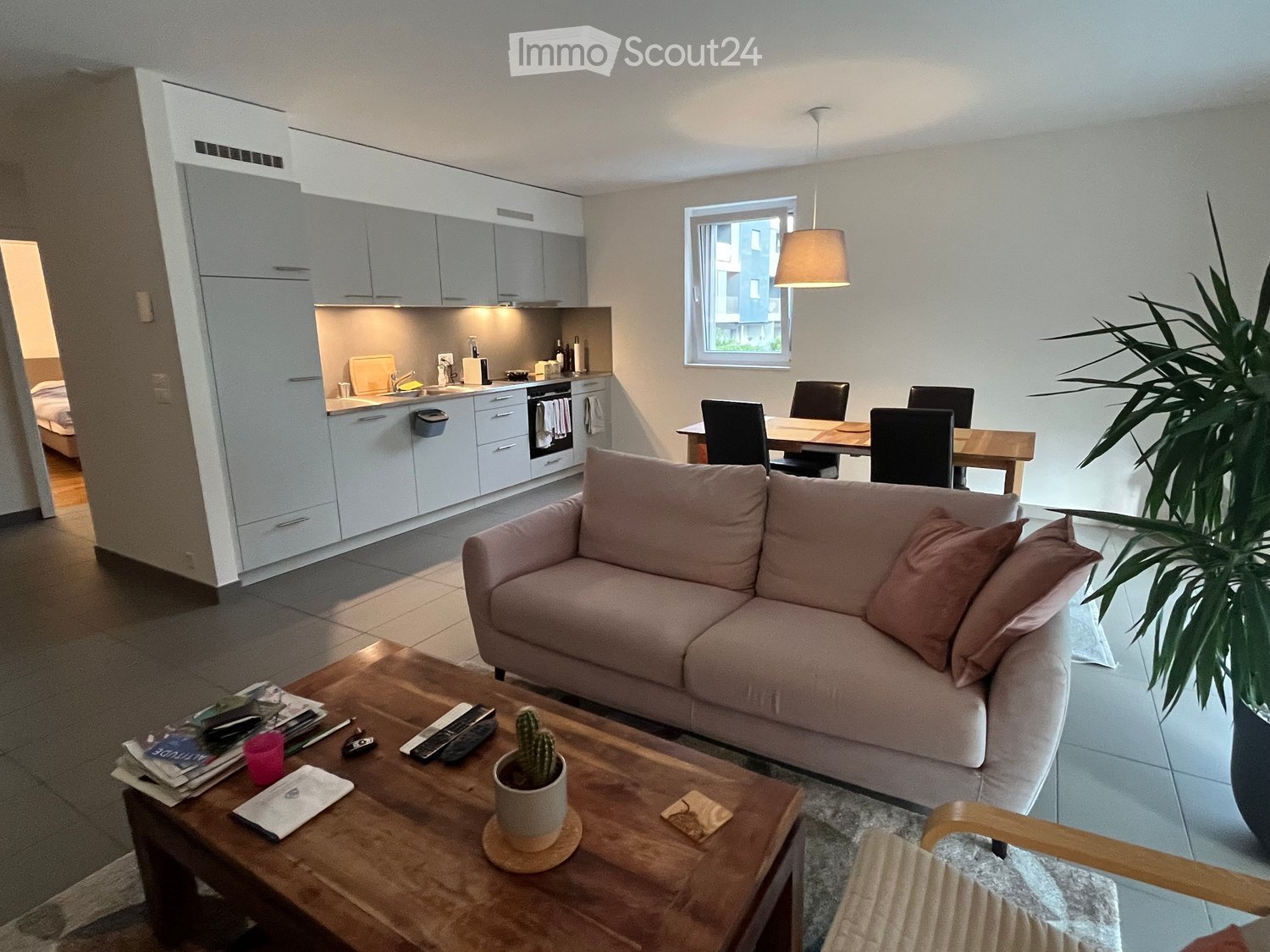 A modern, open-concept living and dining area with a light pink sofa, wooden coffee table, dining table with chairs, and a small kitchen area with white cabinets and stainless steel appliances.