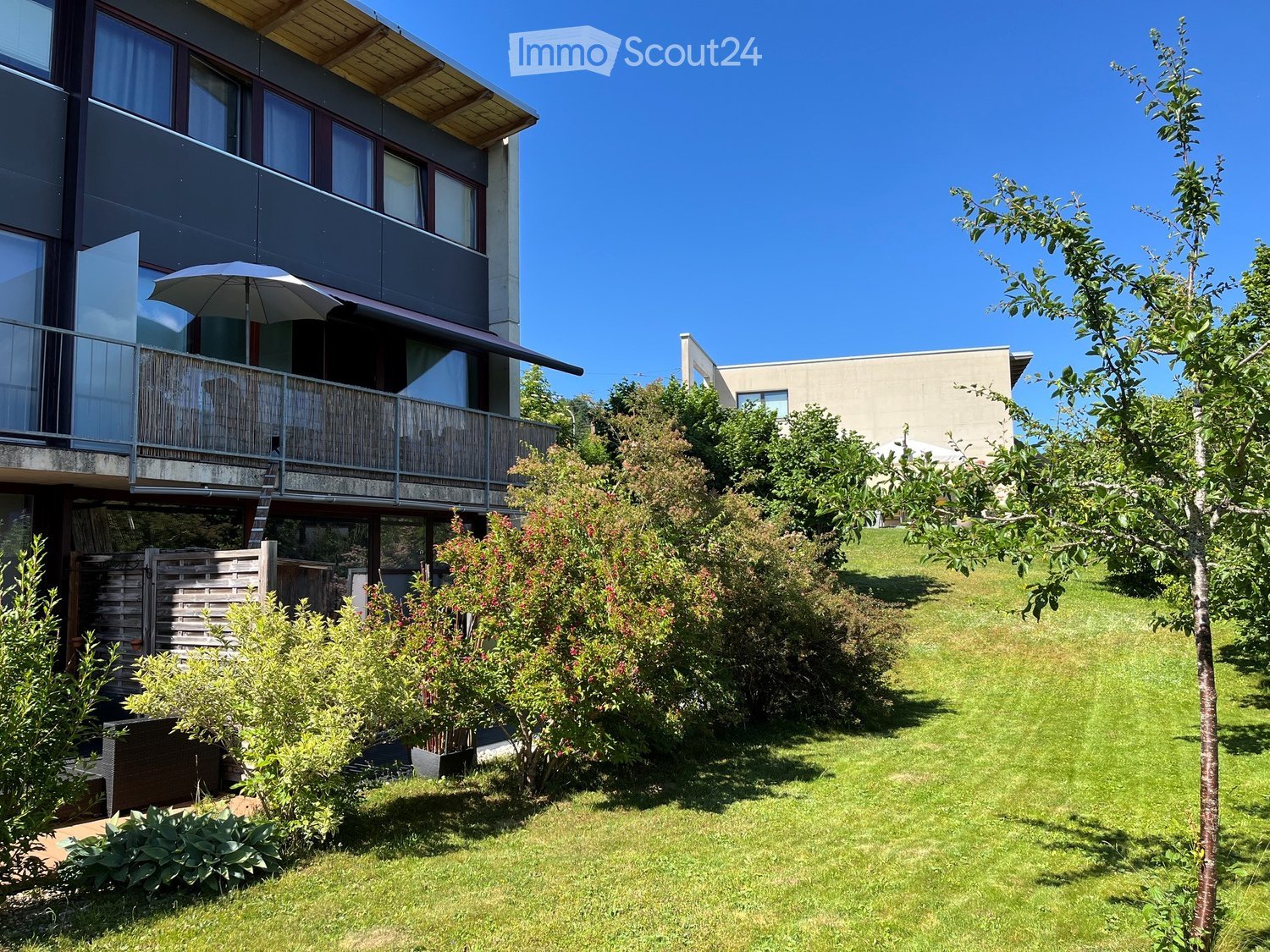 Lush green garden with well-maintained lawn, various shrubs, and trees, featuring a modern multi-story building with balconies and large windows in the background.