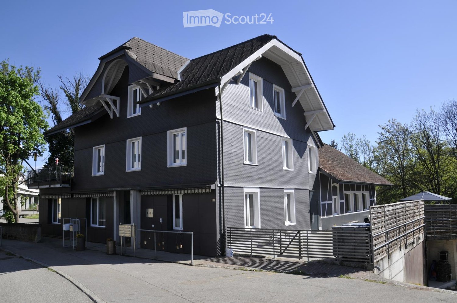 Two-story house with grey brick exterior, white-framed windows, and a balcony on the upper level. The house features a modern design with a sloped roof and a small garden area to the right.