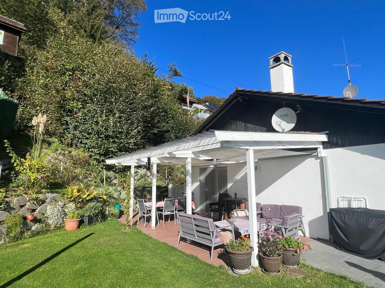 A well-maintained garden with a covered patio area, featuring a sofa, table, chairs, a bench, and several potted plants. The patio is connected to a house with a chimney and satellite dish.