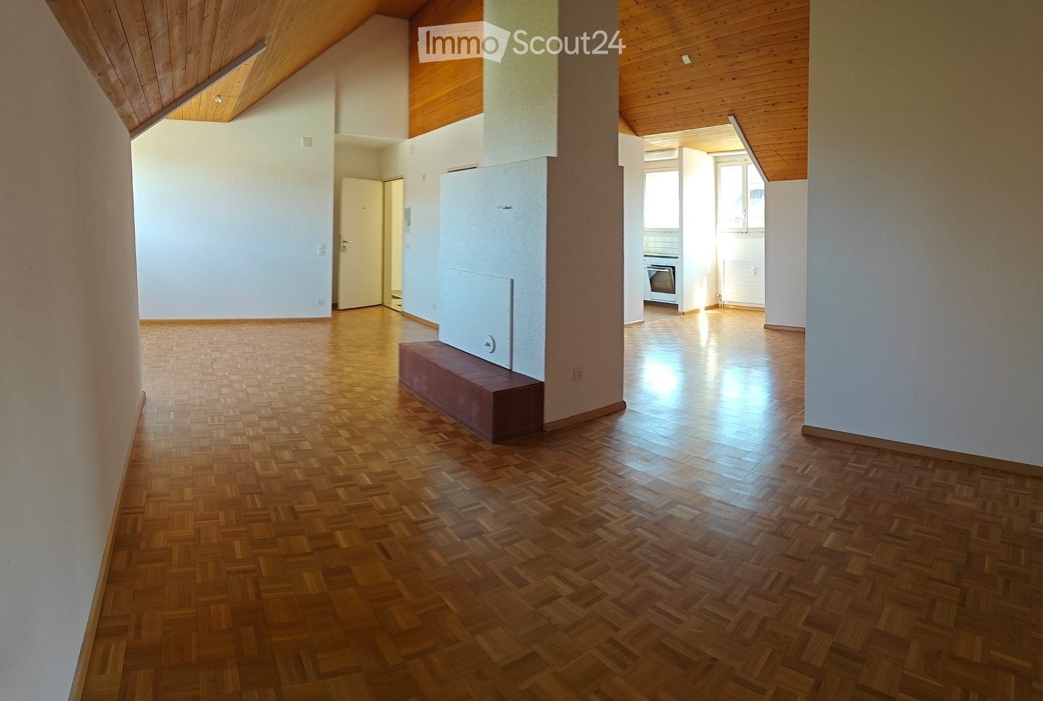 Empty room with wooden flooring, white walls, and a slanted wooden ceiling. A small bench is placed against one wall, and a doorway leads to another room with a visible kitchen area.