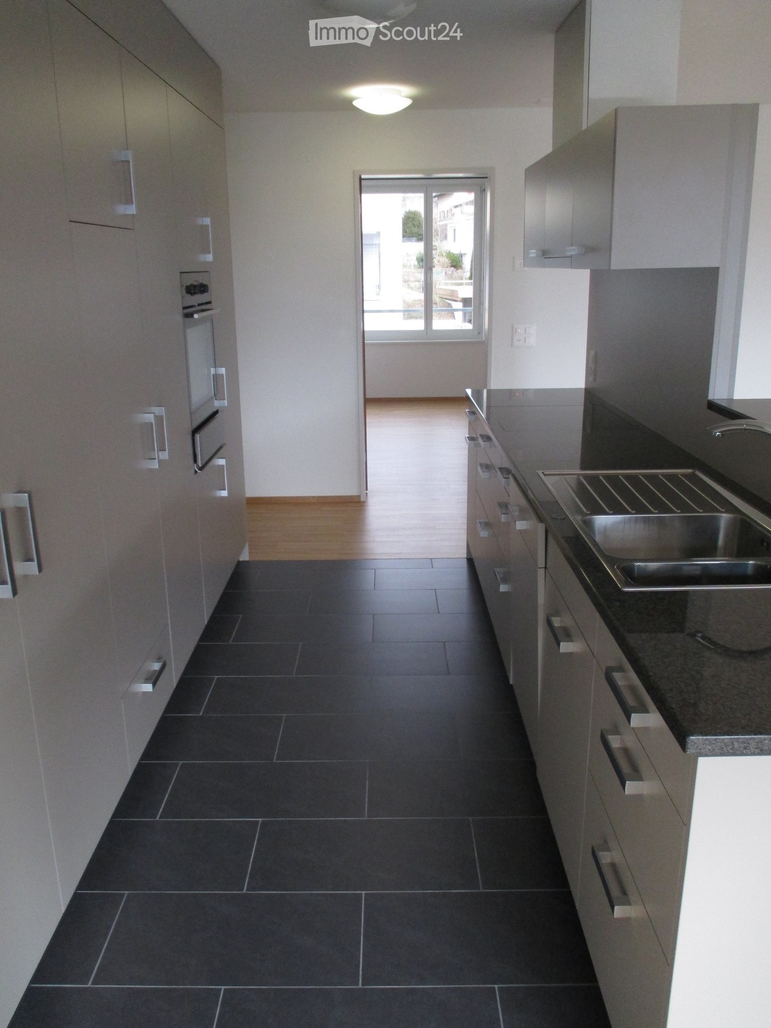 Empty kitchen area with dark tiled floor, light grey cabinets, and a stainless steel sink. The space is well-lit with a ceiling light and has a sliding glass door leading to an outdoor area.