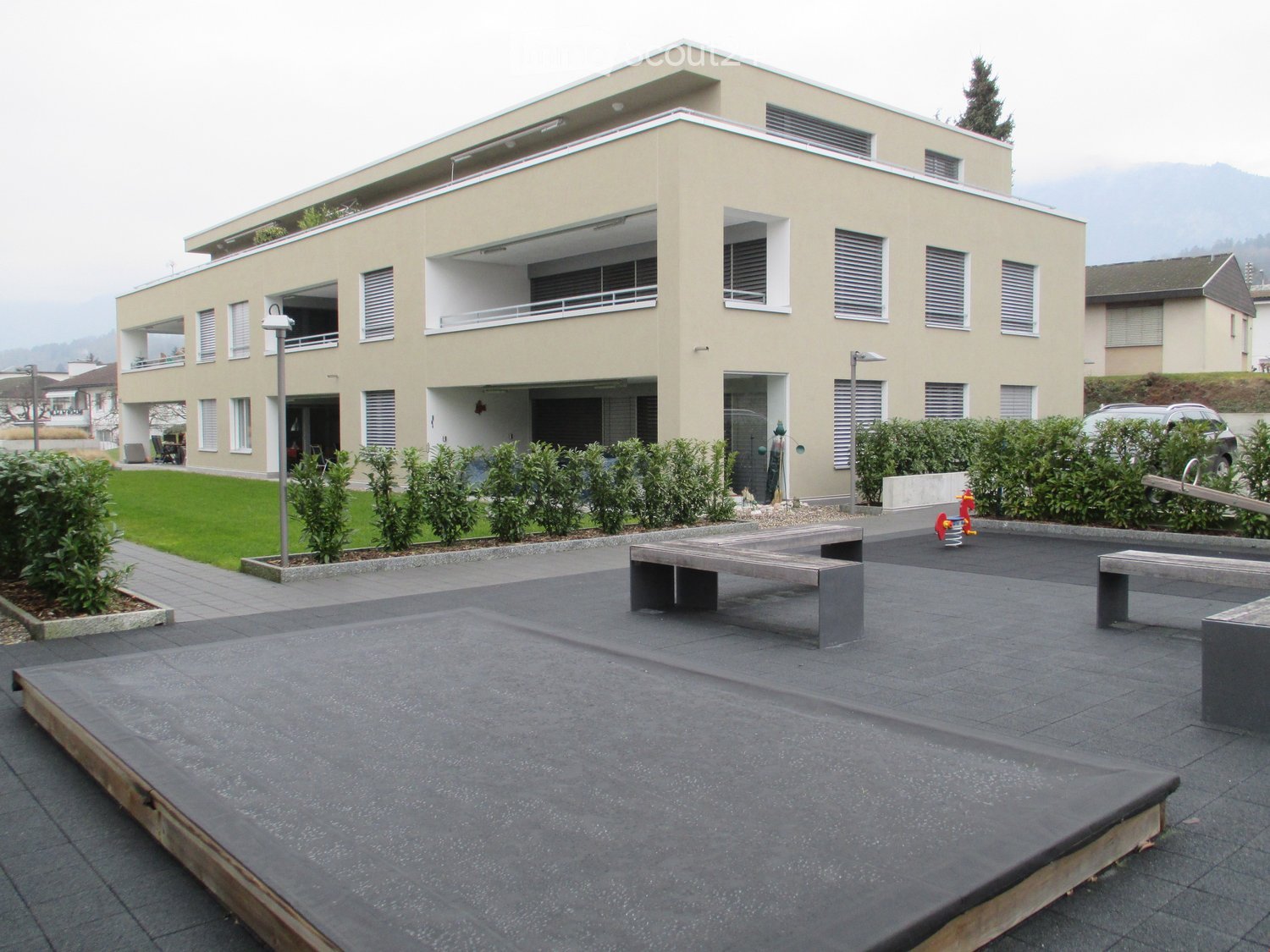 Modern two-story building with beige facade, multiple windows, balconies, and a landscaped courtyard with benches and greenery.