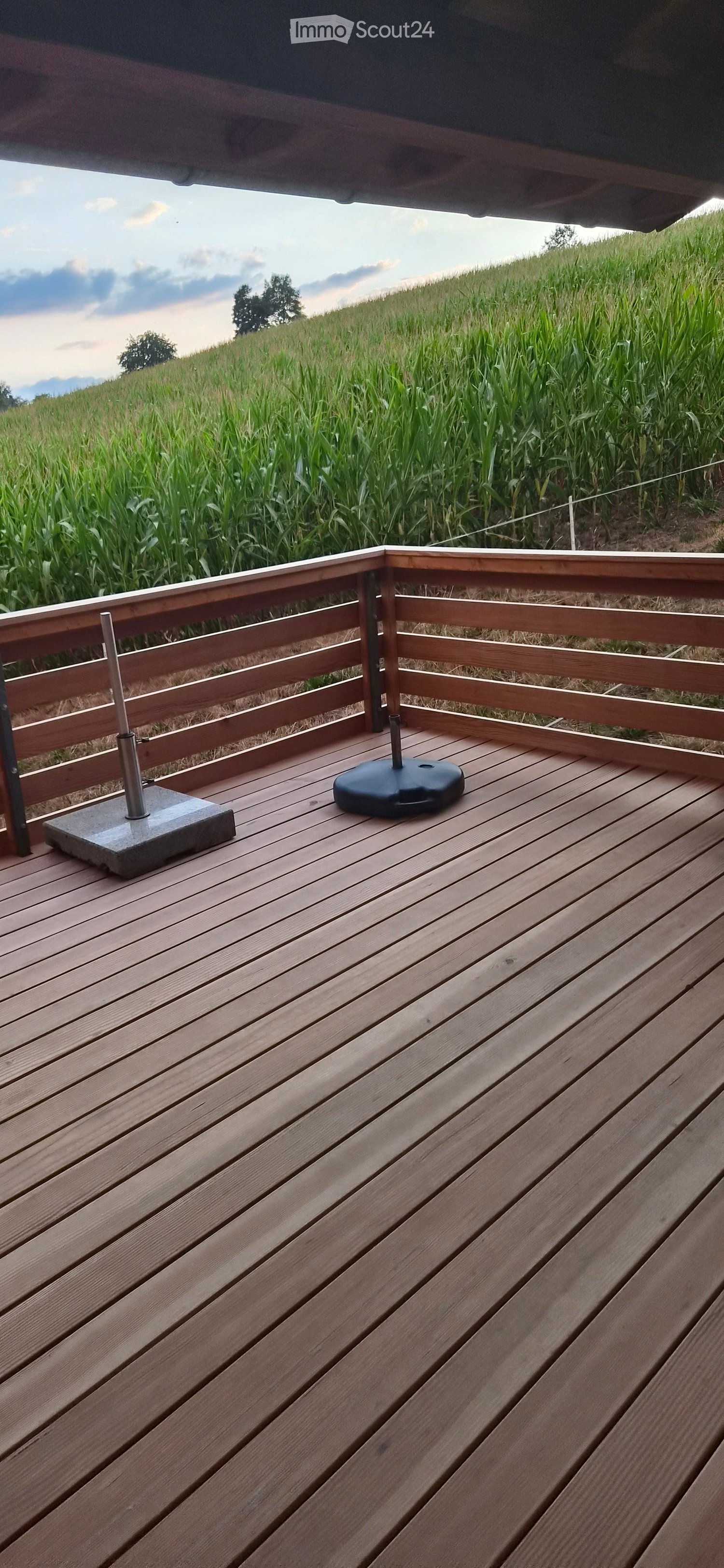 Wooden deck with railing, featuring a concrete base for a parasol and a black parasol base, overlooking a cornfield.