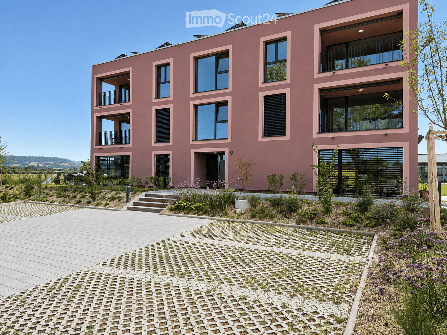 Modern two-story building with pink facade, large windows, and balconies. The entrance features a small garden with plants and flowers, and a paved walkway leads to the building.