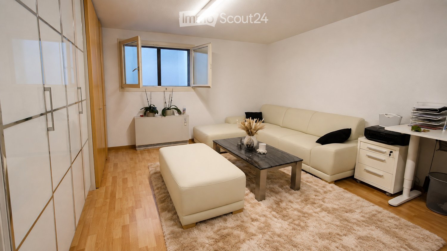 Modern living room with light-colored furniture, including a white sofa, coffee table, ottoman, and a small desk with a chair. The room features wooden flooring, a fluffy white rug, and large windows with open blinds.