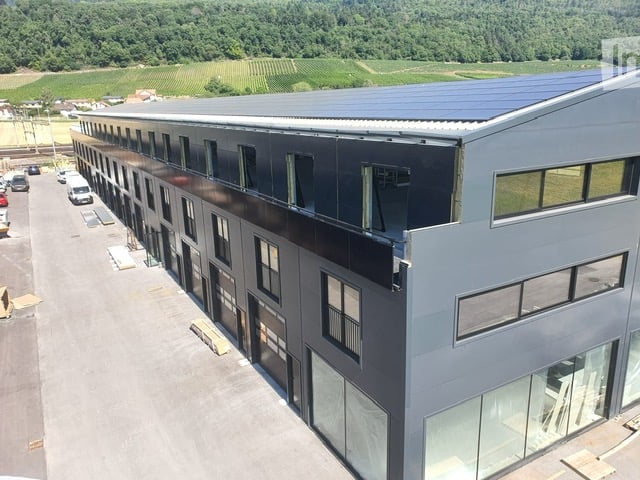 Modern industrial-style building with large windows and a flat roof, surrounded by a parking area with several vehicles.