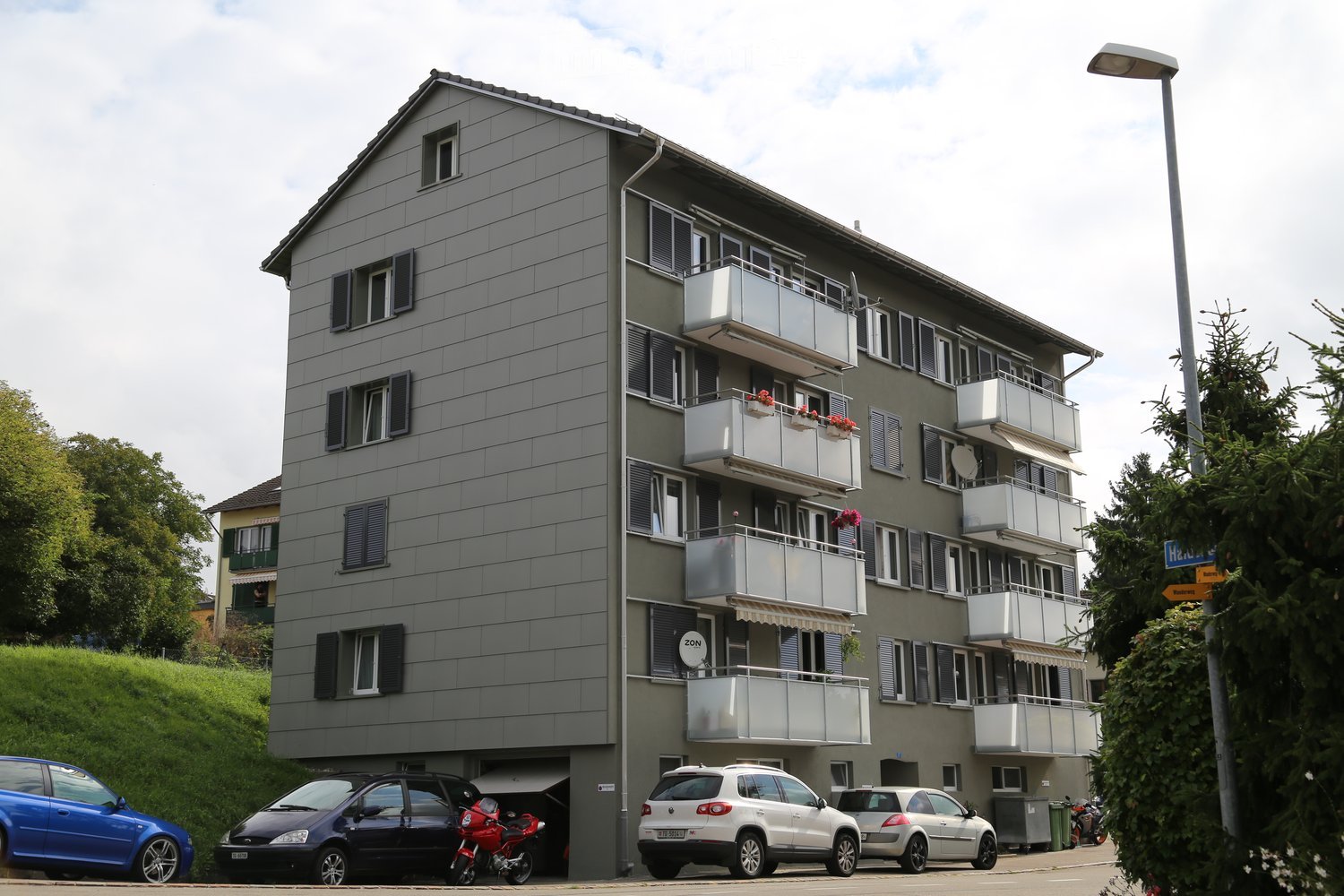 Four-story apartment building with grey facade, multiple balconies, and flower boxes. Several cars and a motorcycle are parked in front of the building.