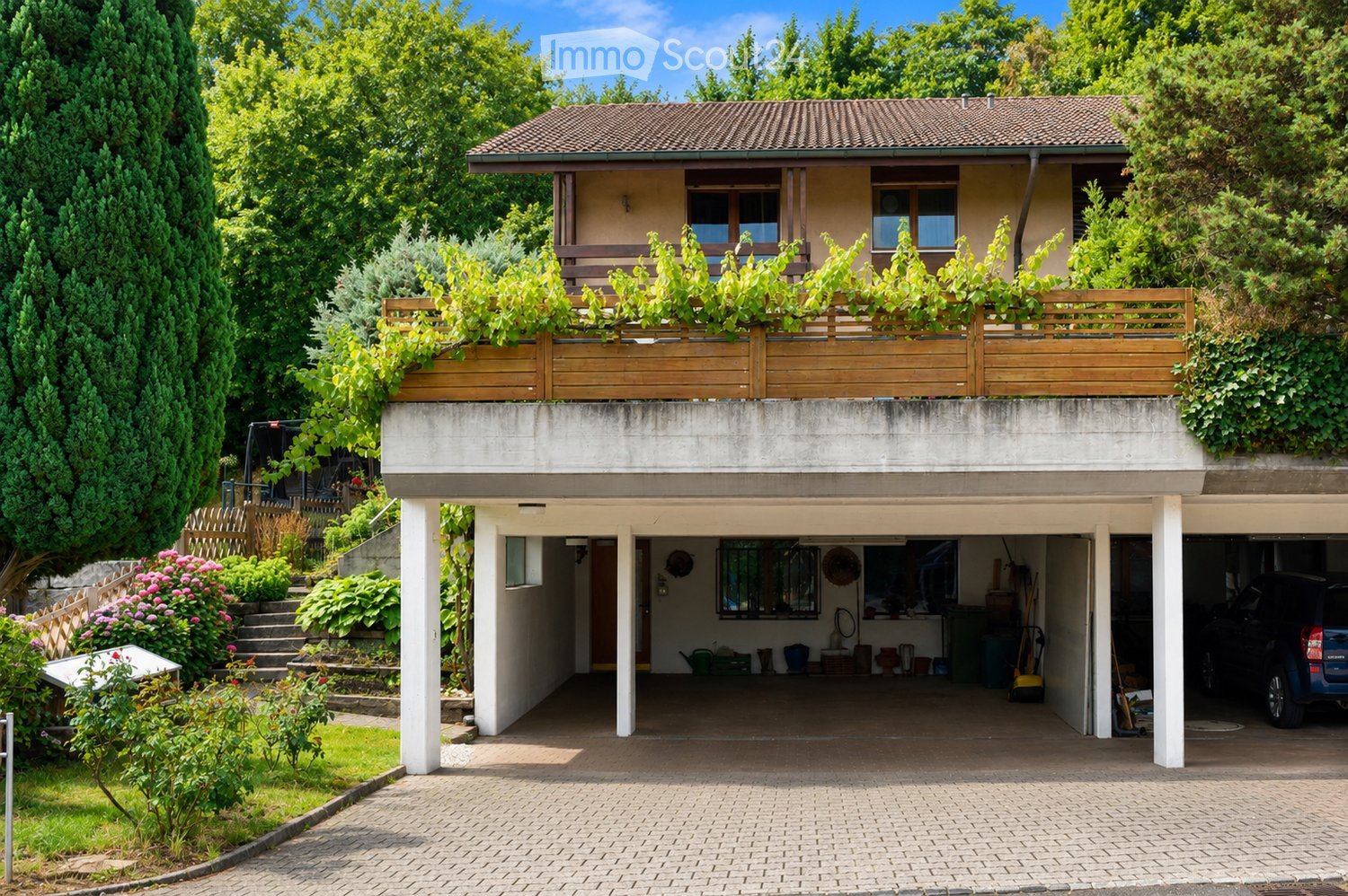 Two-story house with a tiled roof, balcony, garage, and garden area with greenery and flowers.