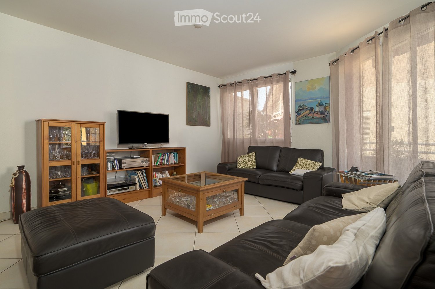 Modern living room with white walls, tiled floor, large windows with curtains, two black leather sofas, a wooden coffee table, an ottoman, a wooden cabinet with glass doors, a bookshelf, and a television on a stand.
