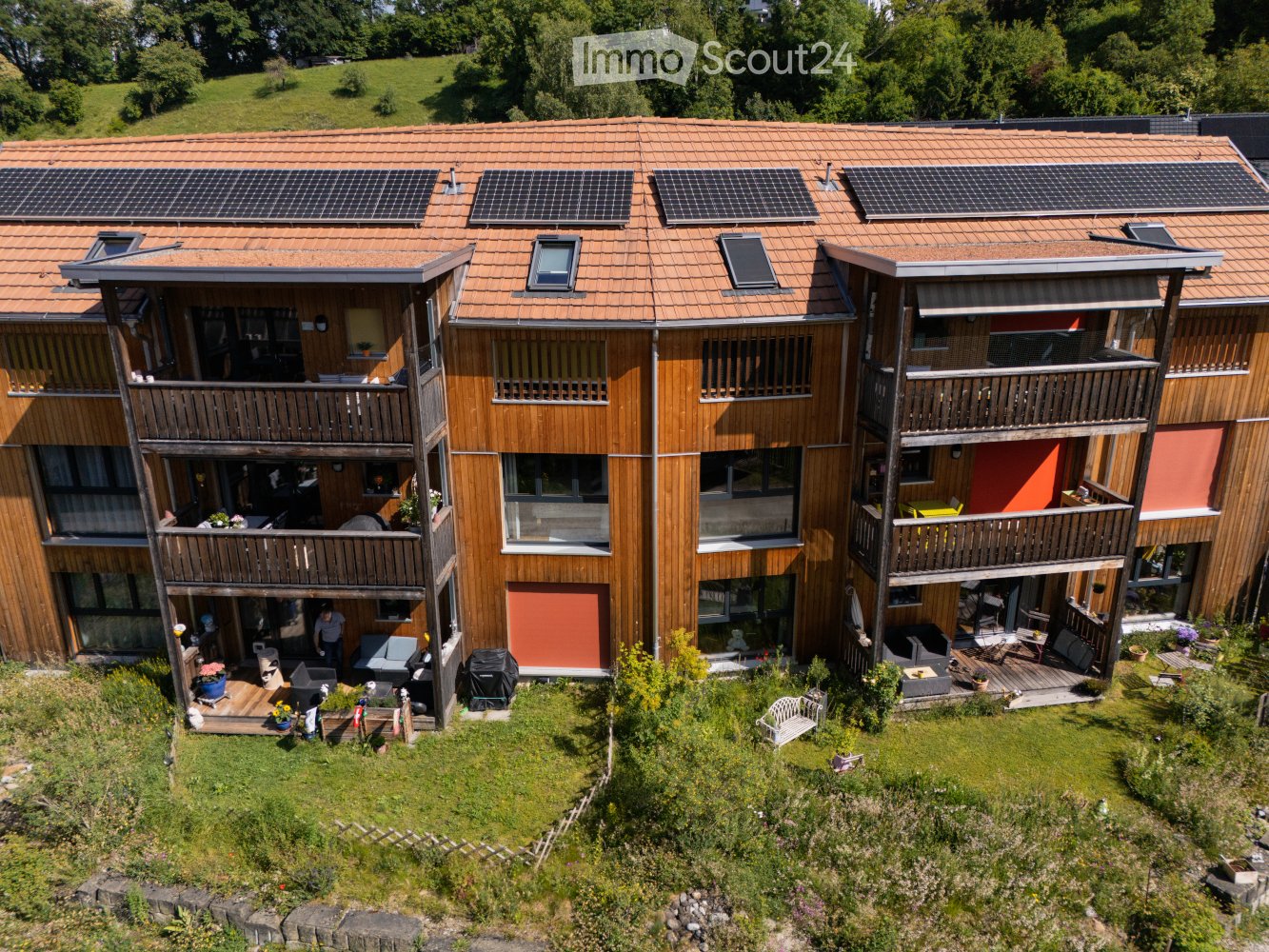 Modern multi-story building with wooden balconies, solar panels on the roof, and a landscaped garden area.