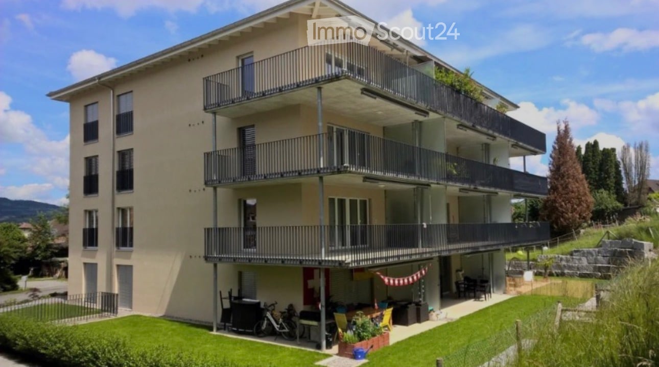 Modern three-story apartment building with multiple balconies, surrounded by greenery and a small garden area with outdoor furniture.