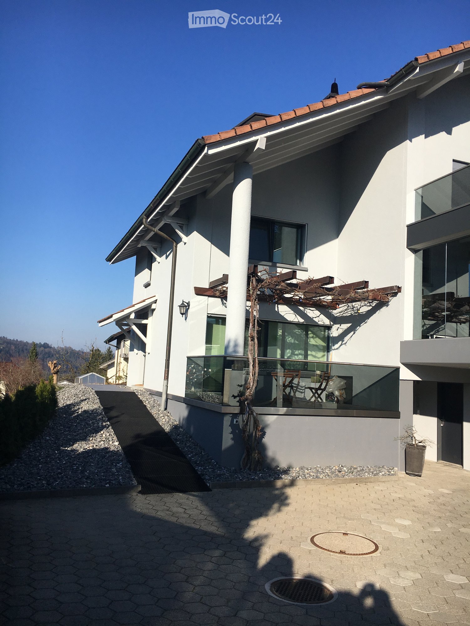 Modern two-story house with white exterior, large windows, and a balcony with glass railing. Features include a raised ground floor, a small garden area, and a paved driveway.