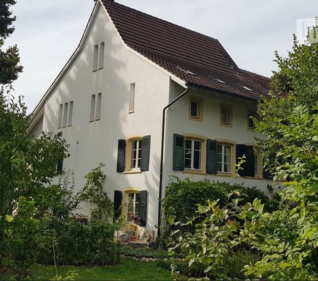 Two-story white house with a brown tiled roof, multiple windows with shutters, and a well-maintained garden in front.