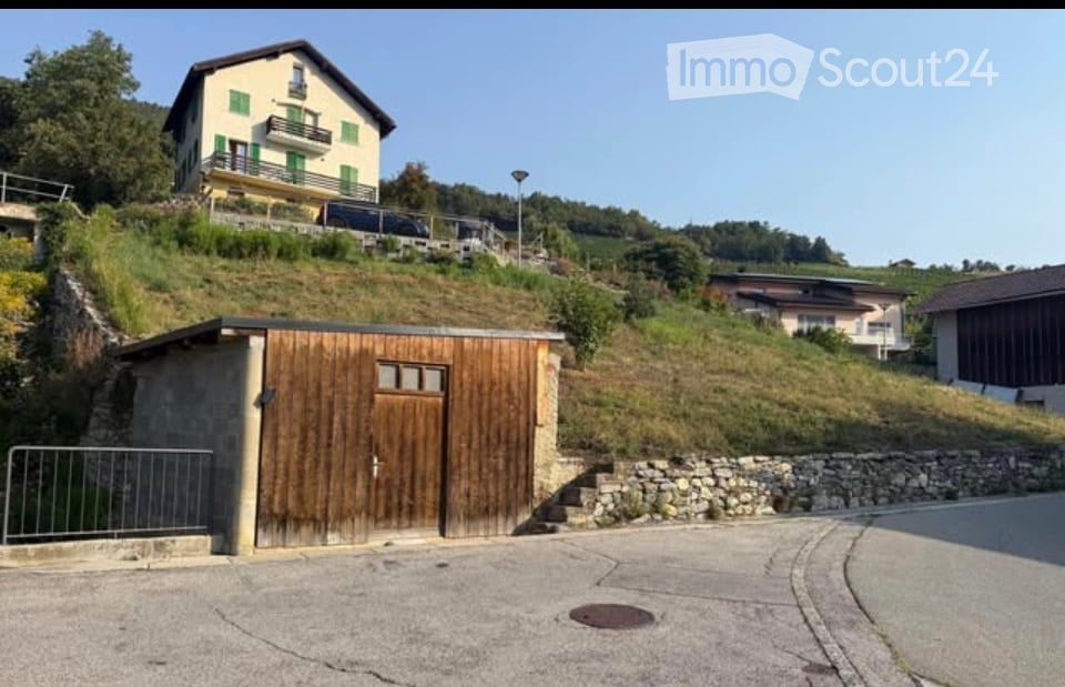 A two-story house with green shutters and balconies sits on a hillside, with a wooden garage at the base of the slope.