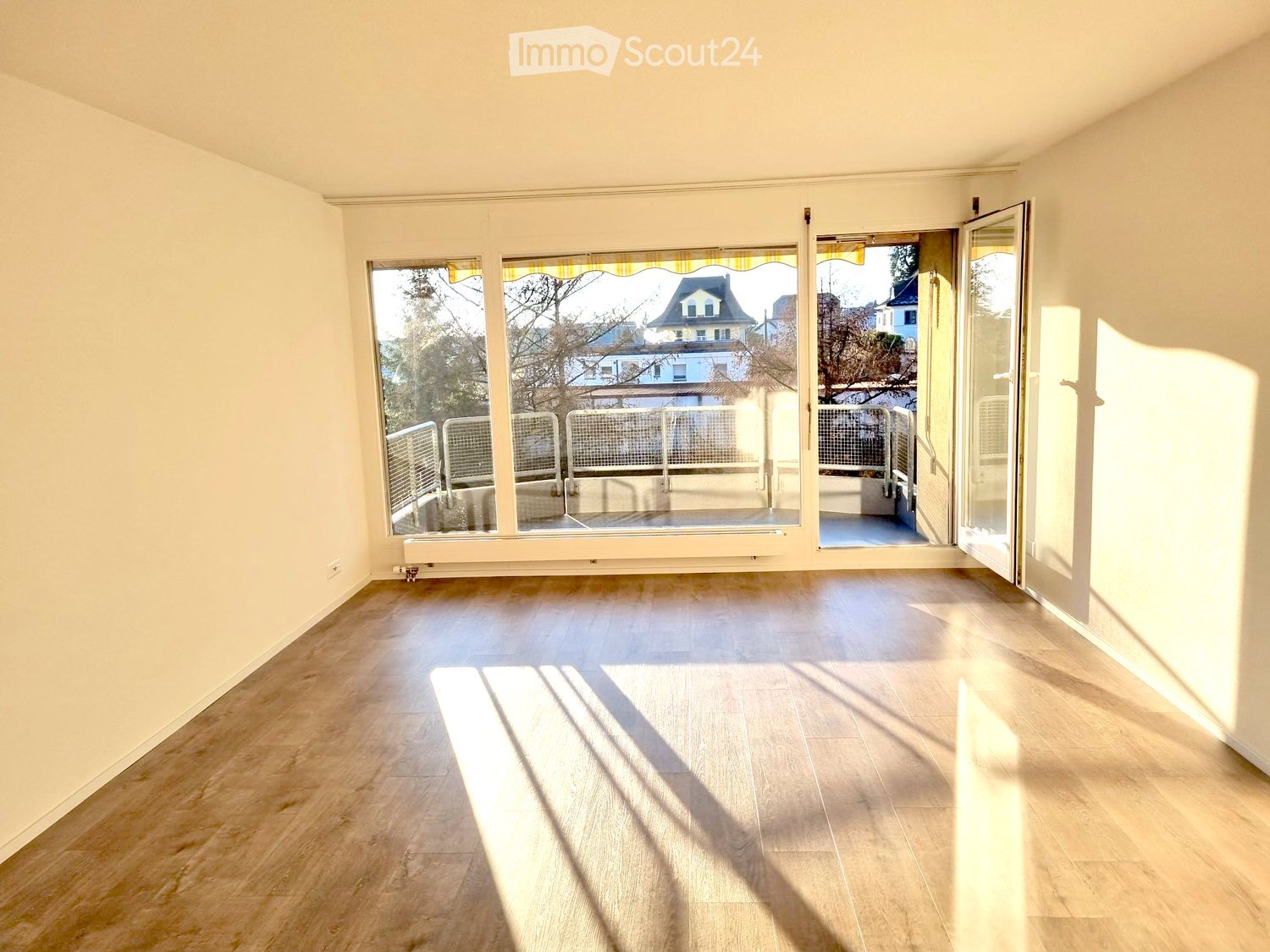 Empty room with light wooden flooring, large windows, and an open sliding door leading to a balcony area.