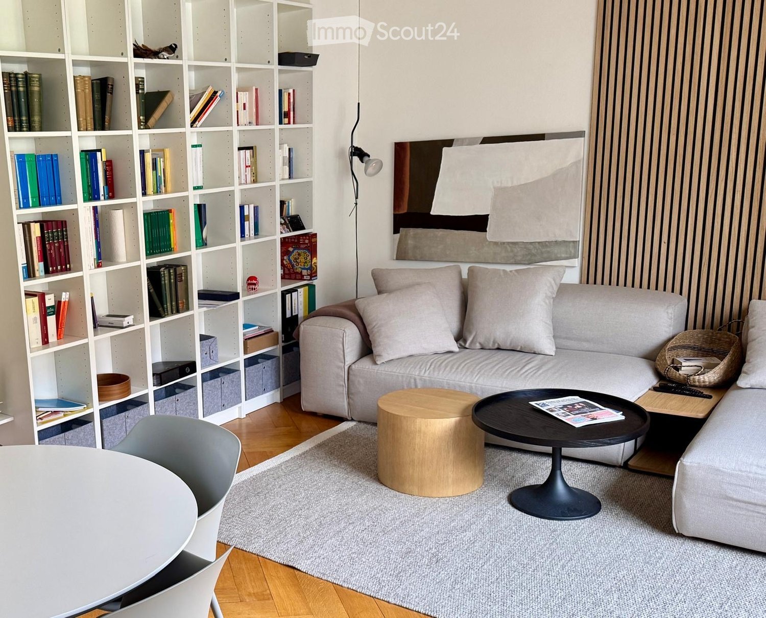 Modern living room with a grey sofa, wooden coffee table, round wooden stool, white dining chair, large bookshelf filled with books, and a grey rug on the floor.