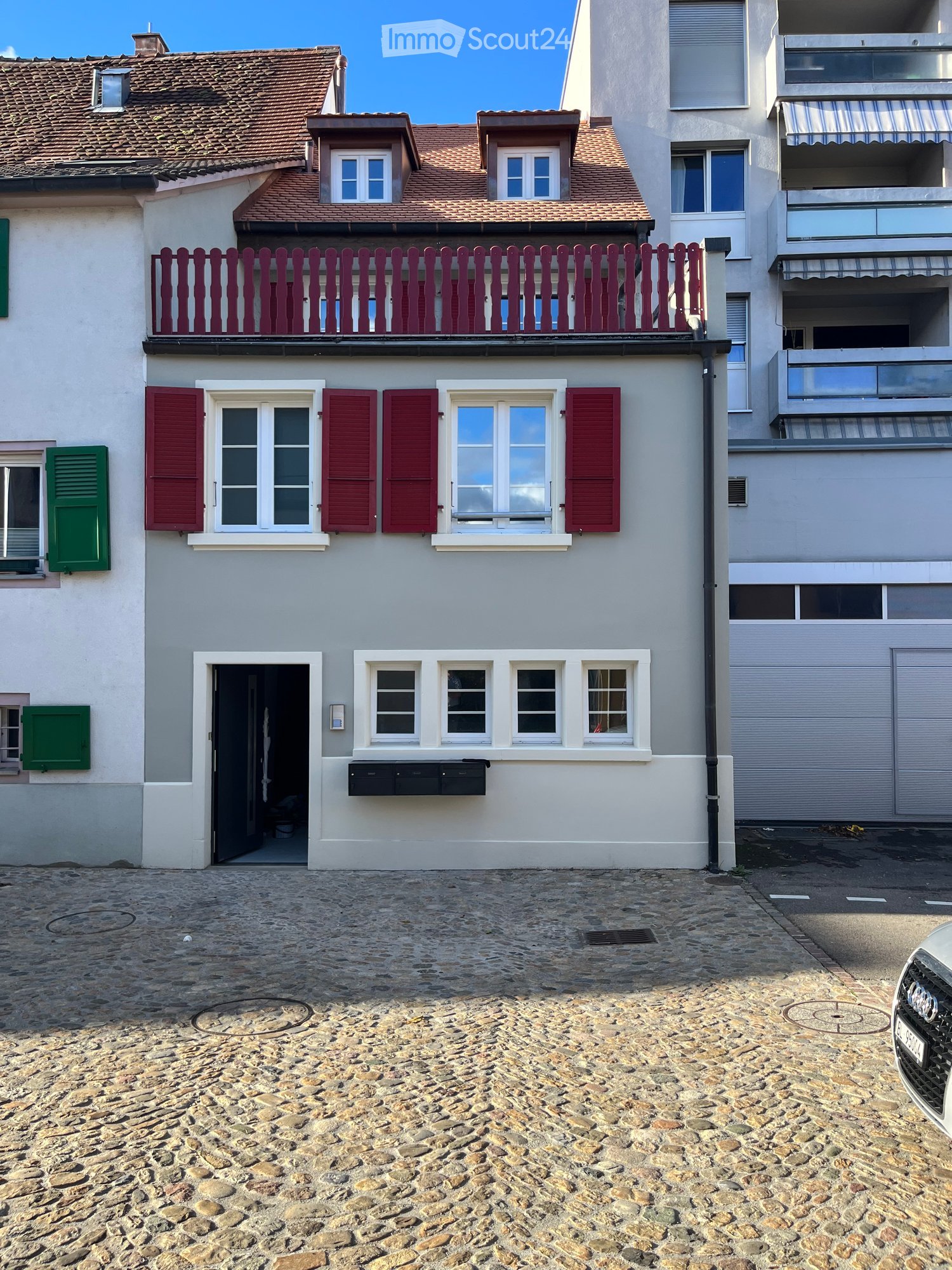 Two-story house with light gray walls, red shutters, and a red balcony railing. The ground floor has a black door and several windows, while the upper floor features additional windows and a small balcony. The roof is tiled with brown tiles and has dormer