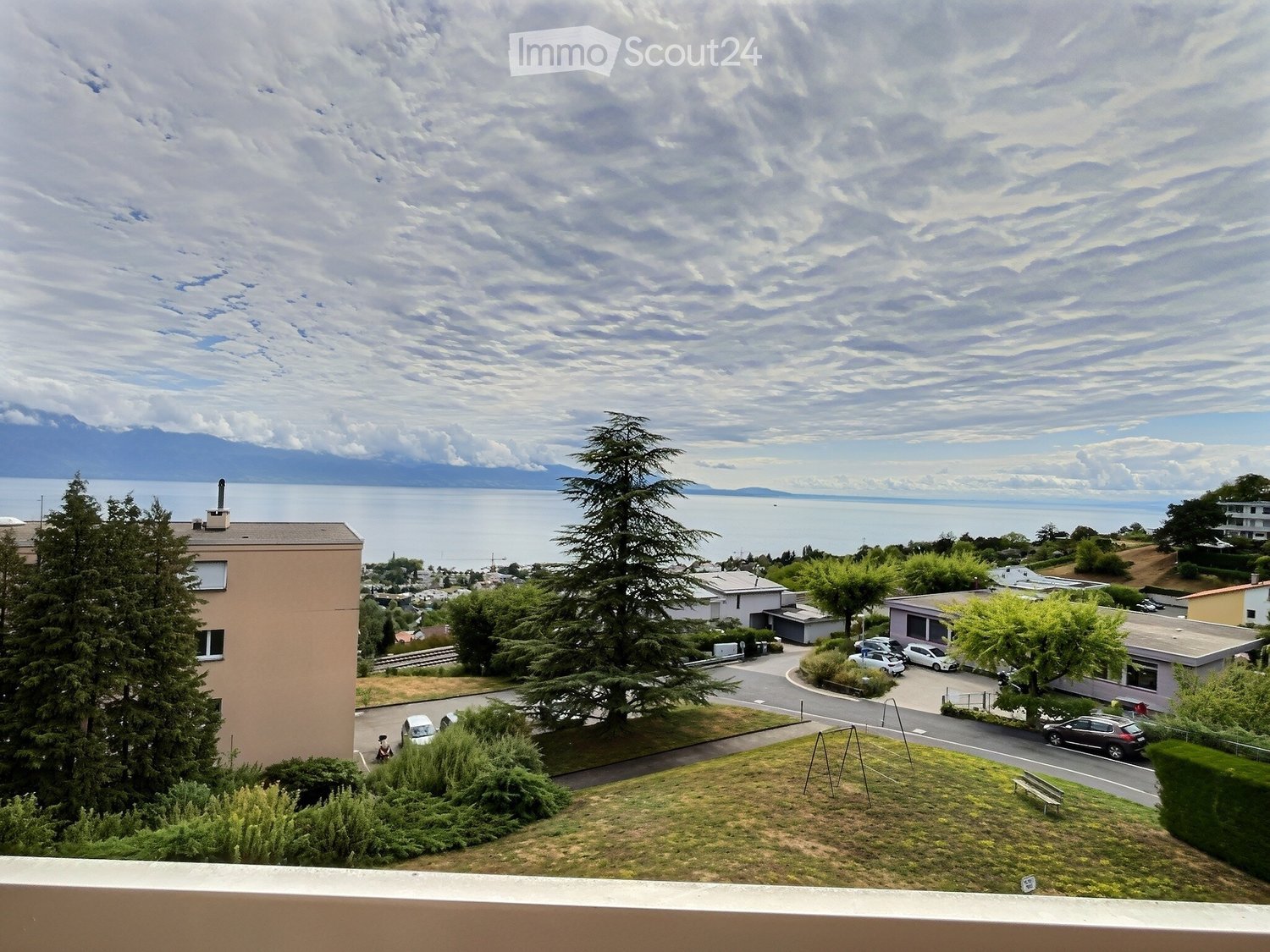 View from a balcony showing a grassy area, trees, parked cars, and a scenic view of a lake and distant mountains.