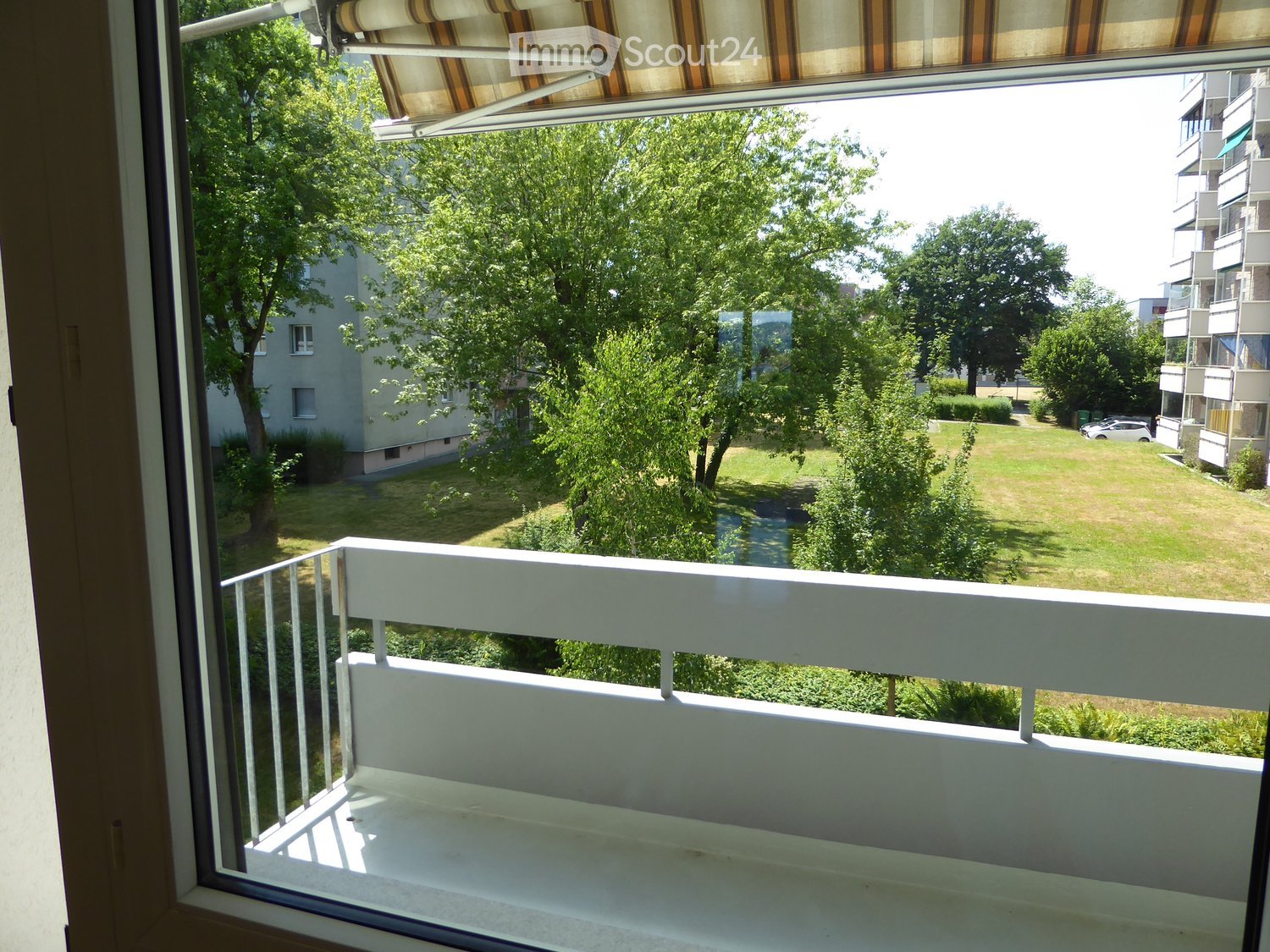 Balcony with white railing, overlooking a green park area with trees and a building in the background.