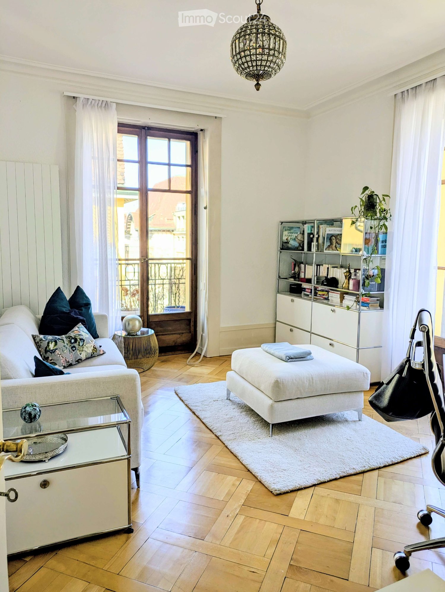 Elegant living room with a white sofa, ottoman, glass coffee table, bookshelf, and a chandelier hanging from the ceiling. The room features wooden flooring and large windows with sheer curtains, leading to a balcony.