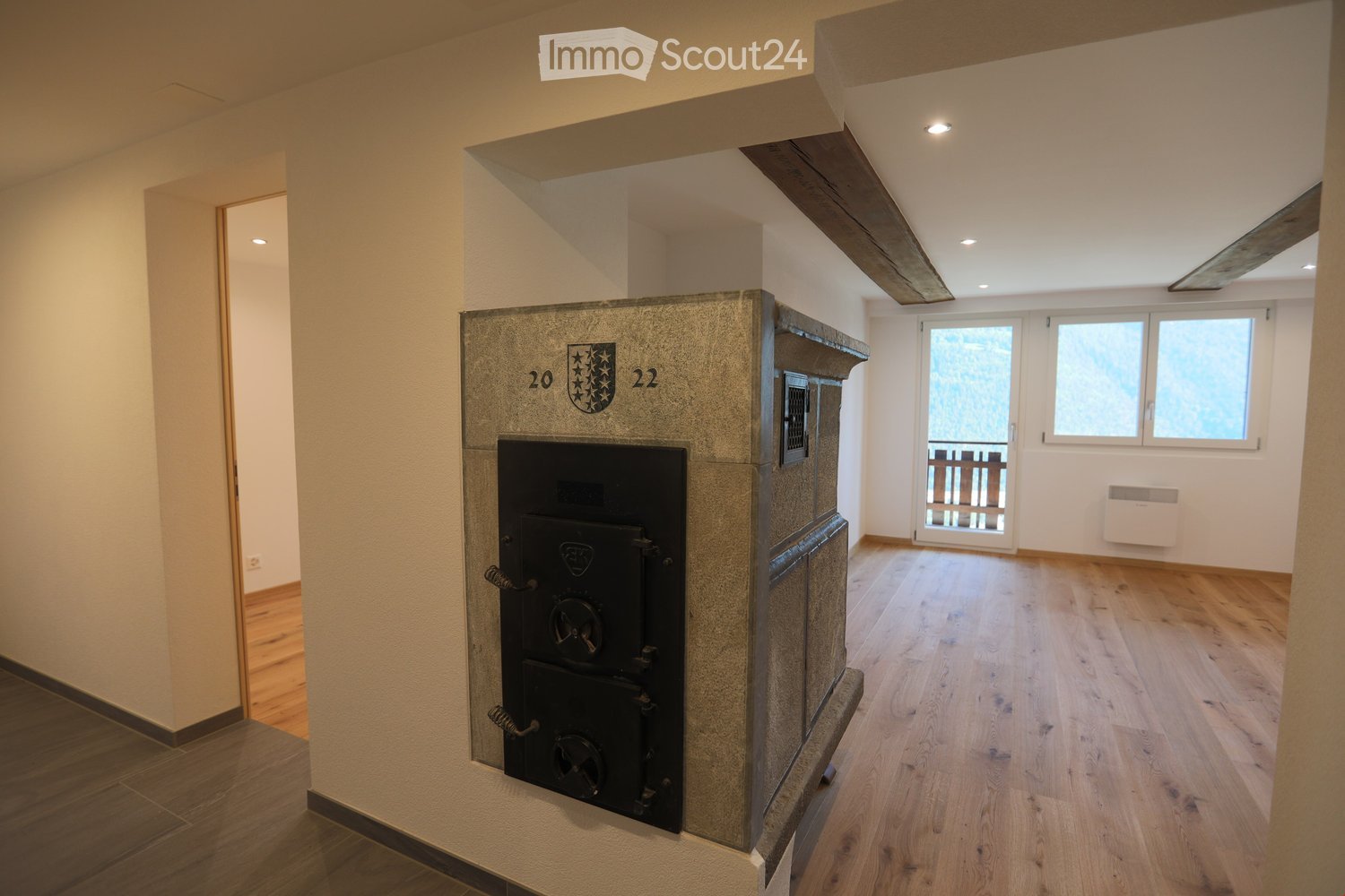 Empty room with wooden flooring, exposed wooden beams on the ceiling, a fireplace with a decorative plaque, and large windows with a view of the outdoors.